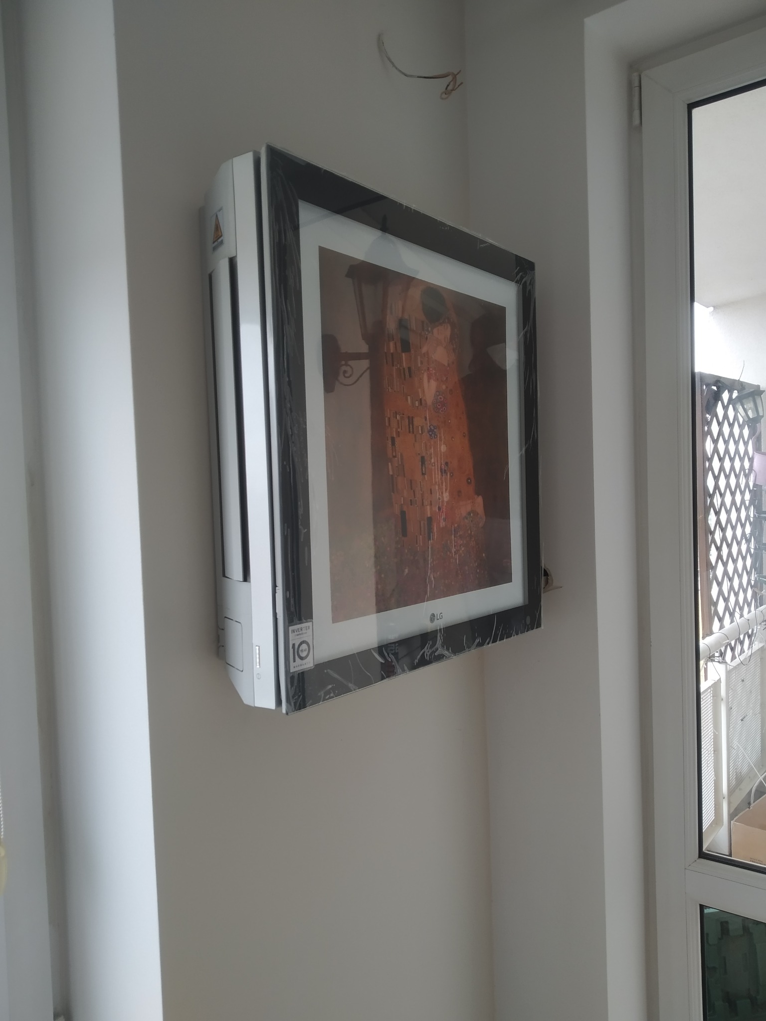 Klimatyzator LG Artcool Gallery z reprodukcją obrazu 'Pocałunek' Gustava Klimta, jeszcze owinięty folią ochronną, zamontowany na białej ścianie w mieszkaniu.