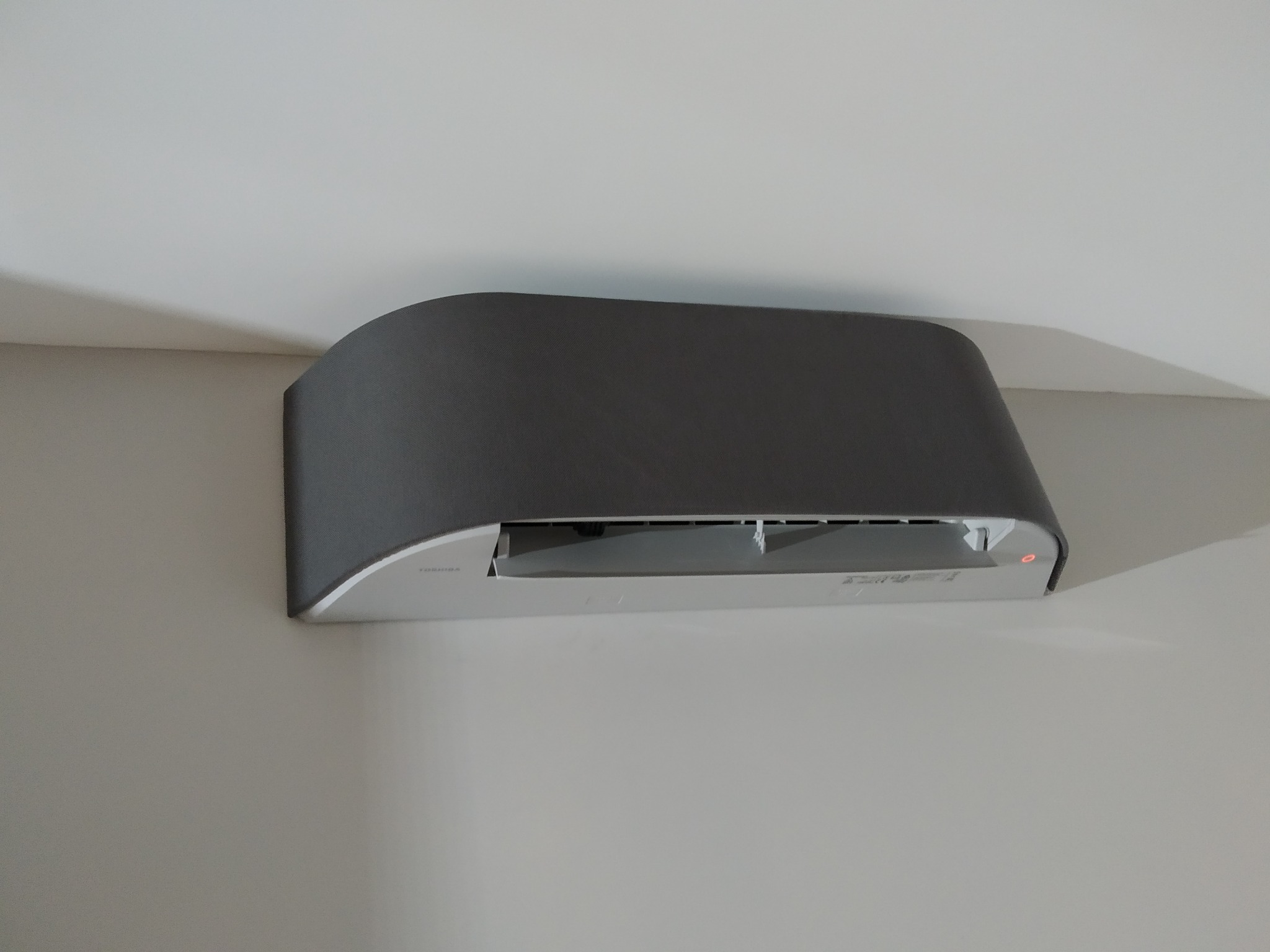 Szary klimatyzator Toshiba zamontowany na białej ścianie, widoczna dioda sygnalizacyjna i subtelne oświetlenie sufitowe.