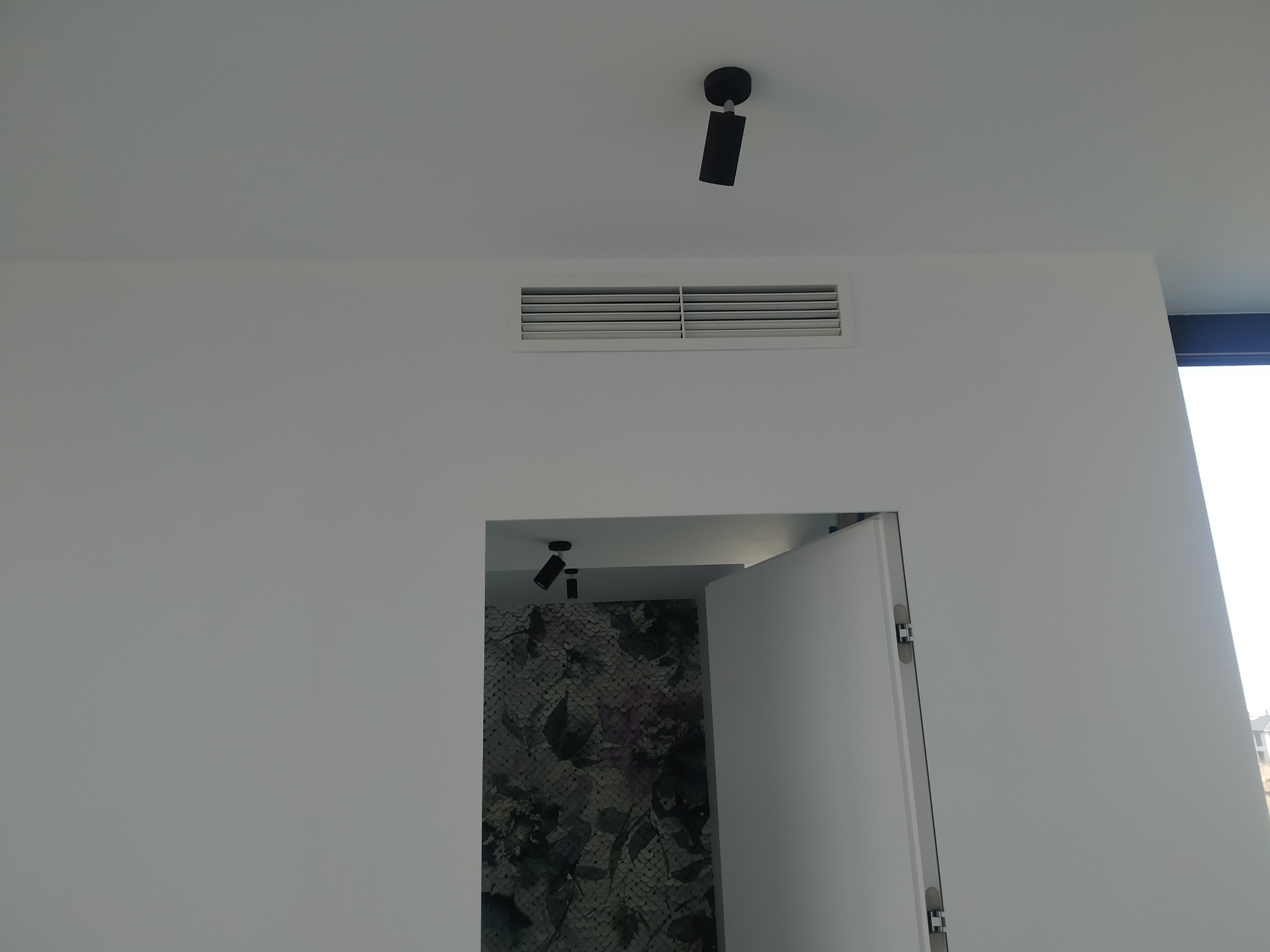 Biała ściana z zamontowaną kratką wentylacyjną i reflektorem sufitowym, widok na otwarte drzwi do pomieszczenia z tapetą w kwiaty.