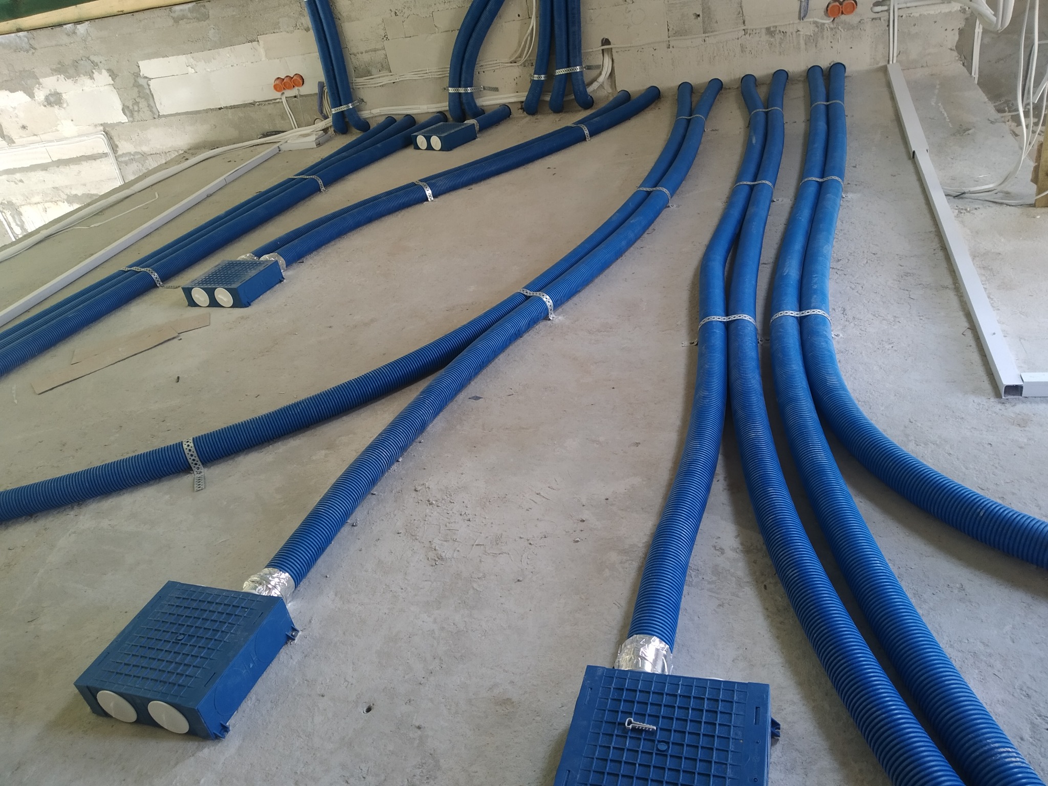 Niebieskie, karbowane rury wentylacyjne ułożone na surowym, betonowym podłożu w trakcie instalacji rekuperacji w budynku. Rury przymocowane metalowymi obejmami, prowadzą do niebieskich skrzynek...