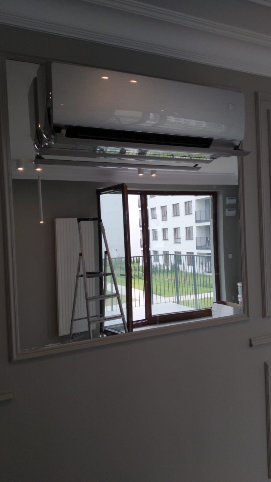Biały klimatyzator ścienny z chromowanymi detalami, widoczny w lustrzanym odbiciu z drabiną i otwartym balkonem w tle.