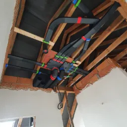 Montaż systemu rekuperacji w trakcie instalacji: czarne, karbowane rury wentylacyjne z kolorowymi oznaczeniami (czerwony, niebieski, zielony) przymocowane do drewnianej konstrukcji stropu, widoczny...