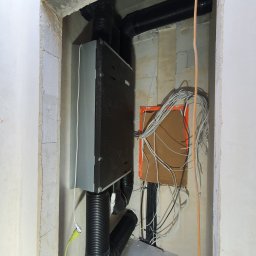 J.P.Rekuperacja Jarosław Pucia - Szary rekuperator z podłączonymi czarnymi rurami wentylacyjnymi w surowym pomieszczeniu, widoczna instalacja elektryczna w pomarańczowej obudowie.