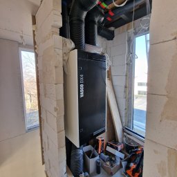 J.P.Rekuperacja Jarosław Pucia - Czarna jednostka rekuperacyjna Vasco DX4 z podłączonymi czarnymi rurami wentylacyjnymi, widoczna w surowym wnętrzu budynku w trakcie budowy, z oknem w tle i narzędziami obok.