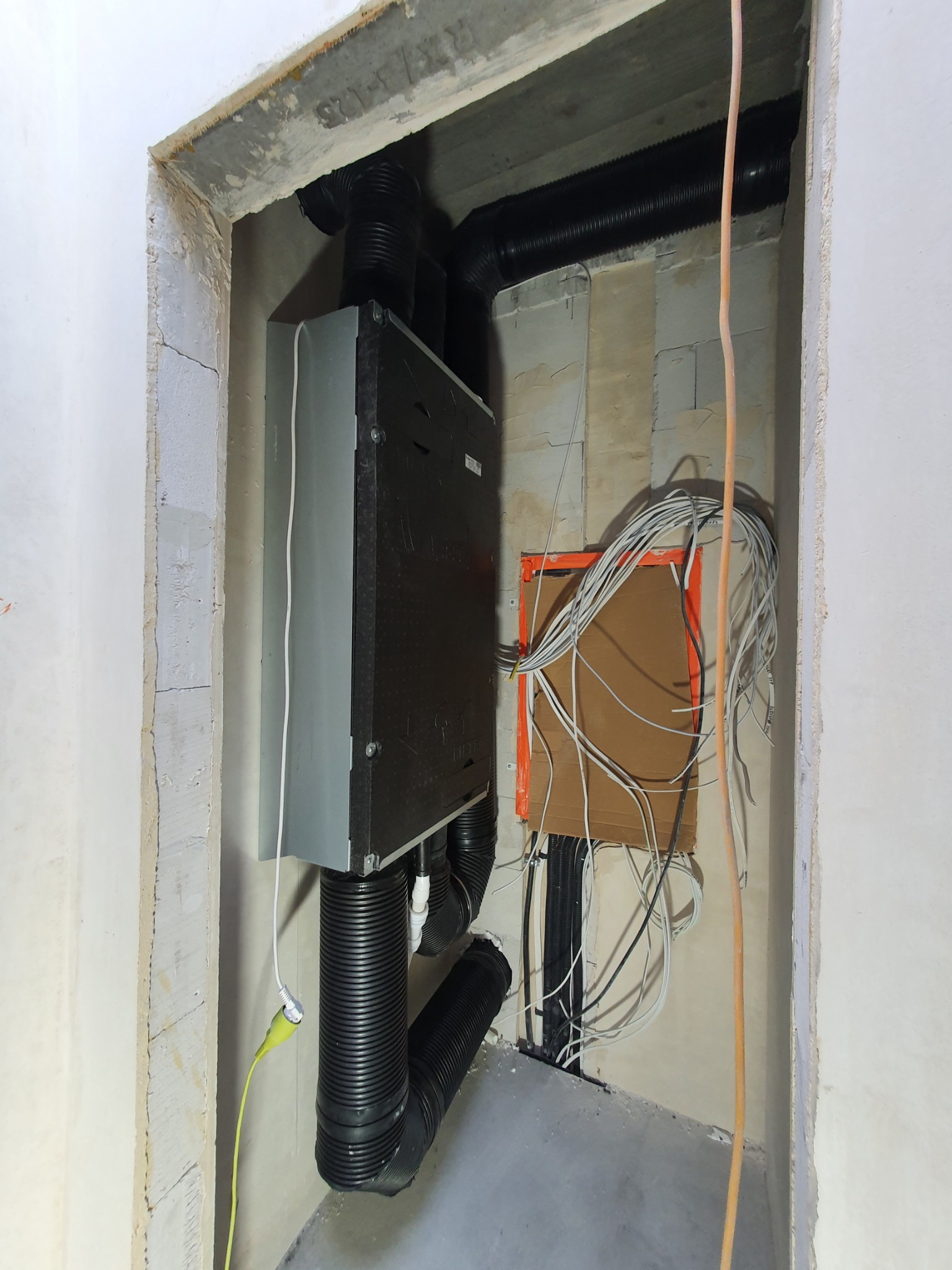 Szary rekuperator z podłączonymi czarnymi rurami wentylacyjnymi w surowym pomieszczeniu, widoczna instalacja elektryczna w pomarańczowej obudowie.