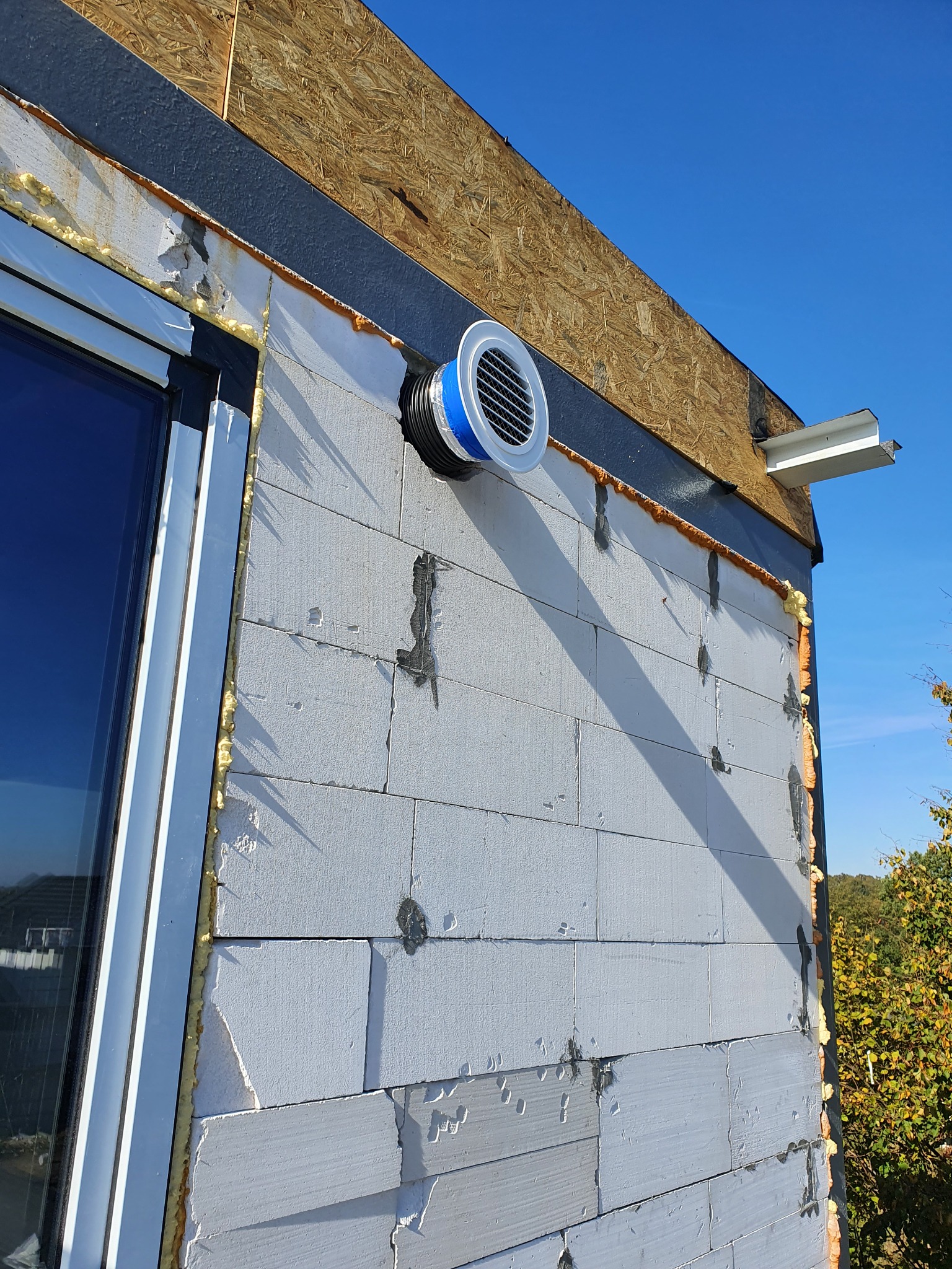 Fasada budynku w trakcie ocieplania, widoczny wlot wentylacyjny z niebieskim akcentem, szara listwa dachowa i fragment okna.