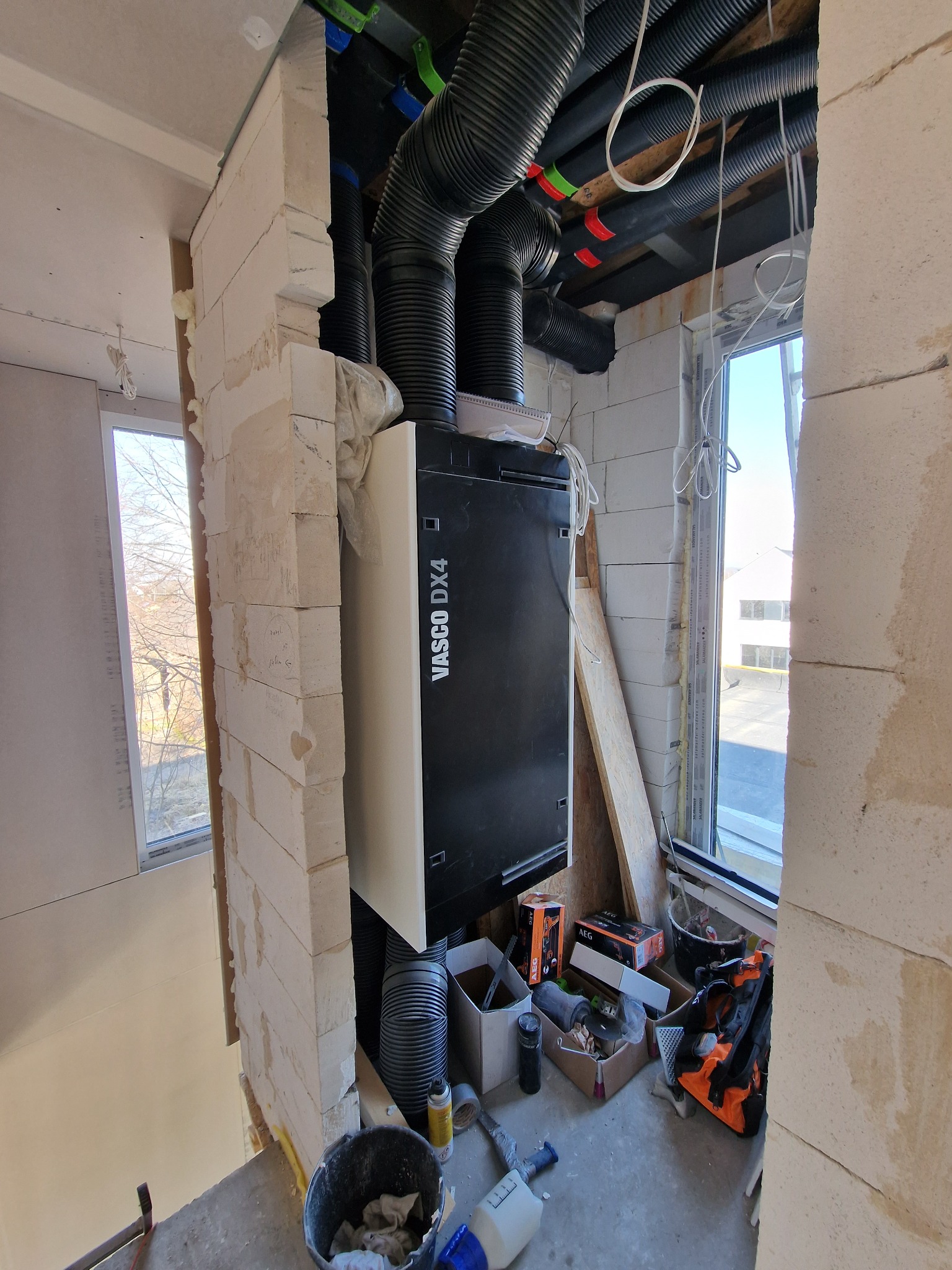 Czarna jednostka rekuperacyjna Vasco DX4 z podłączonymi czarnymi rurami wentylacyjnymi, widoczna w surowym wnętrzu budynku w trakcie budowy, z oknem w tle i narzędziami obok.