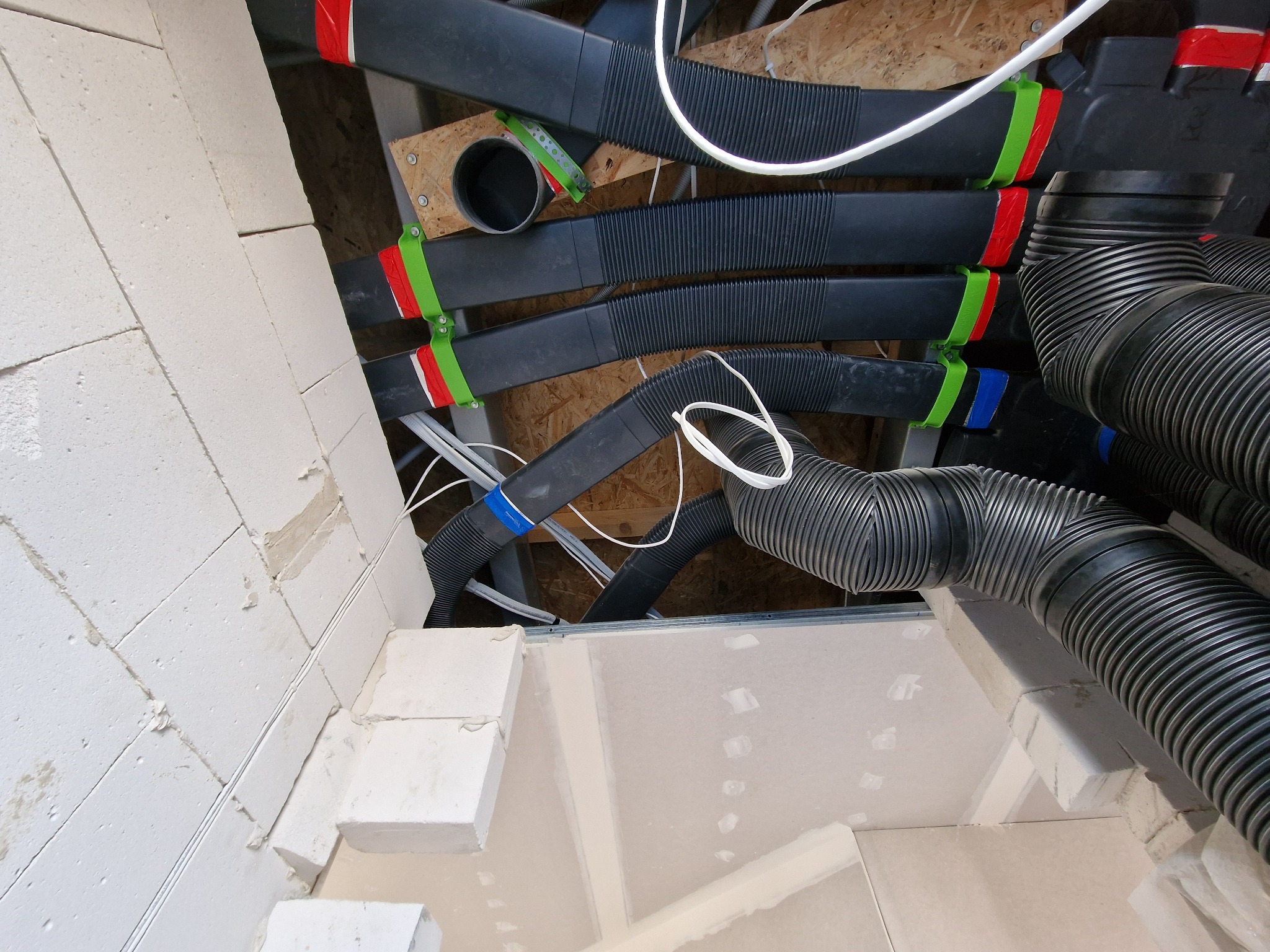 Fragment instalacji wentylacyjnej z czarnymi, karbowanymi rurami przymocowanymi do sufitu za pomocą kolorowych uchwytów (czerwony, zielony, niebieski), widoczne białe przewody i fragment ściany...