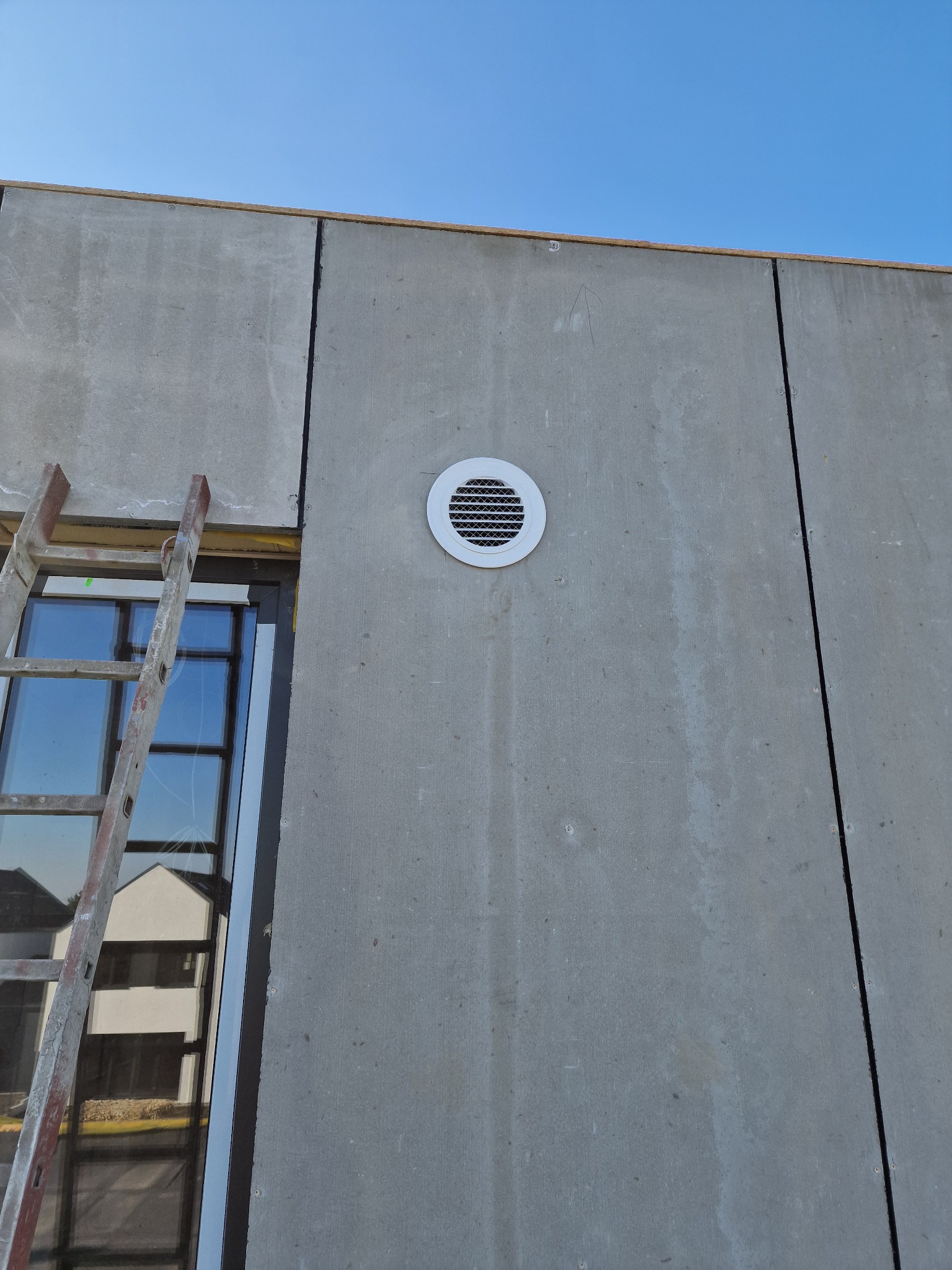 Szara ściana budynku z zamontowanym okrągłym anemostatem wentylacyjnym z białym obramowaniem, obok drabina przystawna oparta o okno.