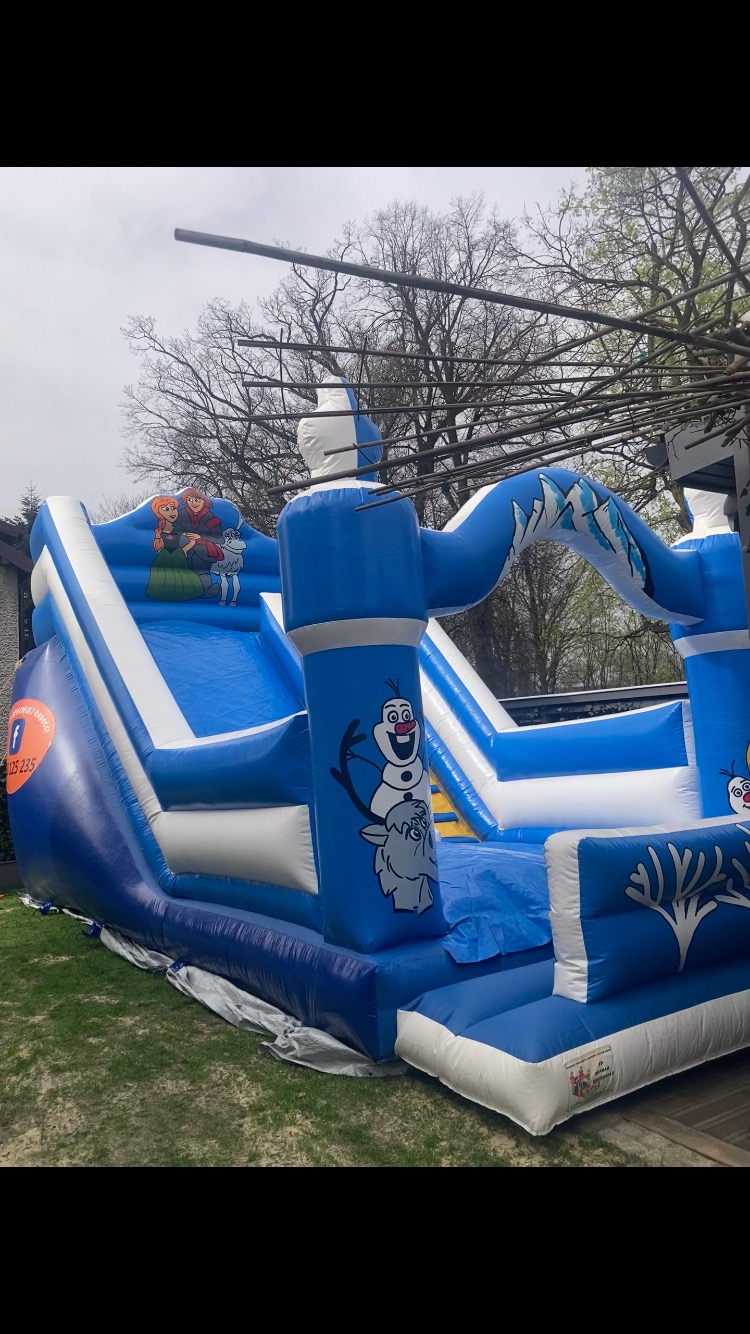 Niebiesko-biały dmuchany zamek ze zjeżdżalnią, ozdobiony motywami z 'Krainy Lodu', ustawiony na trawniku.