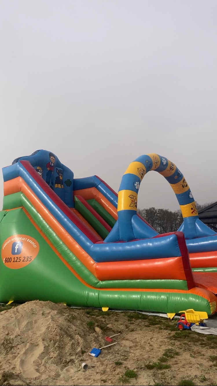 Wielokolorowy dmuchany zamek ze zjeżdżalnią i motywami z bajek, obok zabawka - mała ciężarówka i łopatki w piasku.