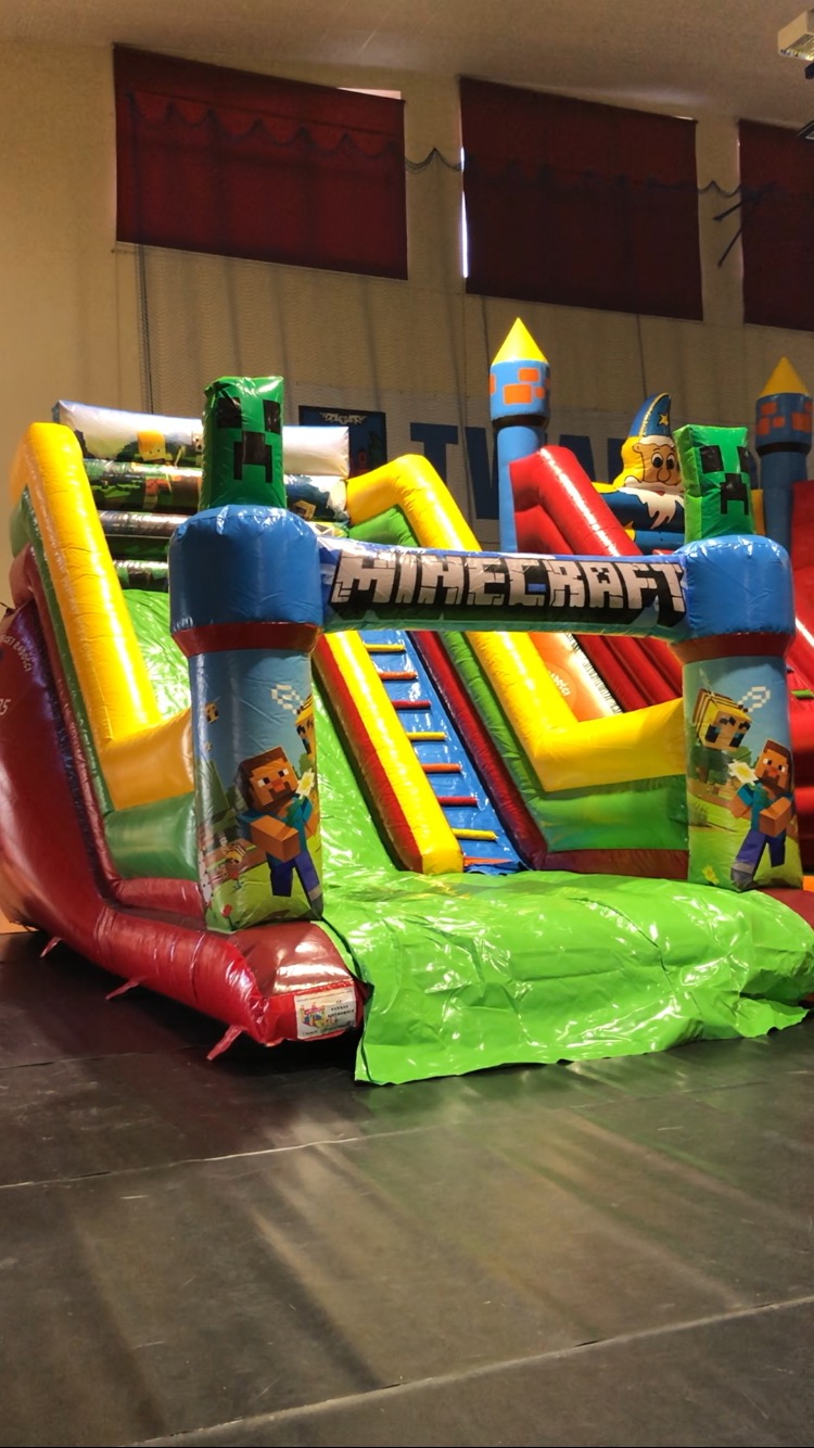 Wielobarwny dmuchany zamek ze zjeżdżalniami i motywem Minecraft, ustawiony wewnątrz pomieszczenia z czerwonymi zasłonami w tle.