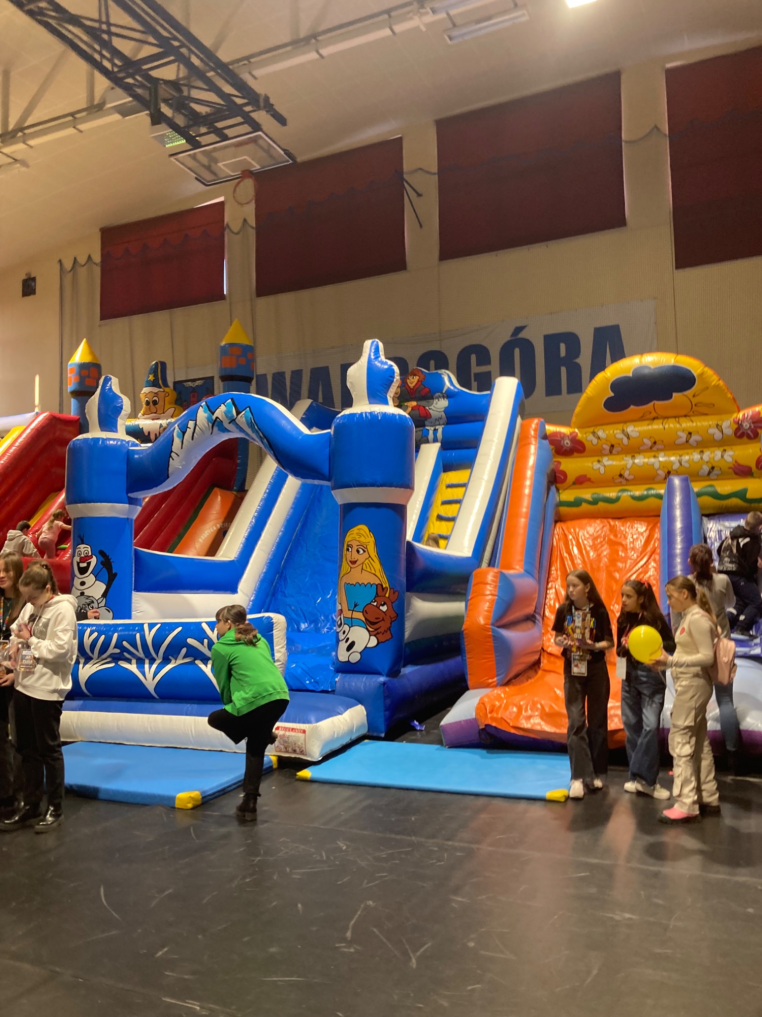 Wnętrze hali z rozstawionymi dmuchanymi zamkami i zjeżdżalniami o tematyce bajkowej, dzieci w tle.