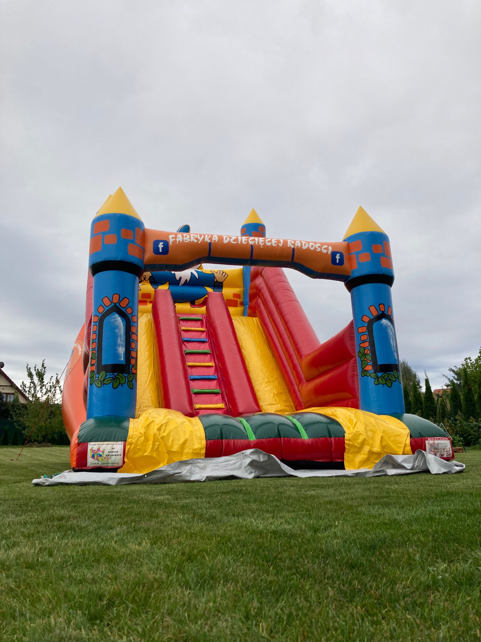 Kolorowy, dmuchany zamek ze zjeżdżalnią na trawniku, z widocznym logotypem 'Fabryka Dziecięcej Radości' i ikonami Facebooka, otoczony zielenią.