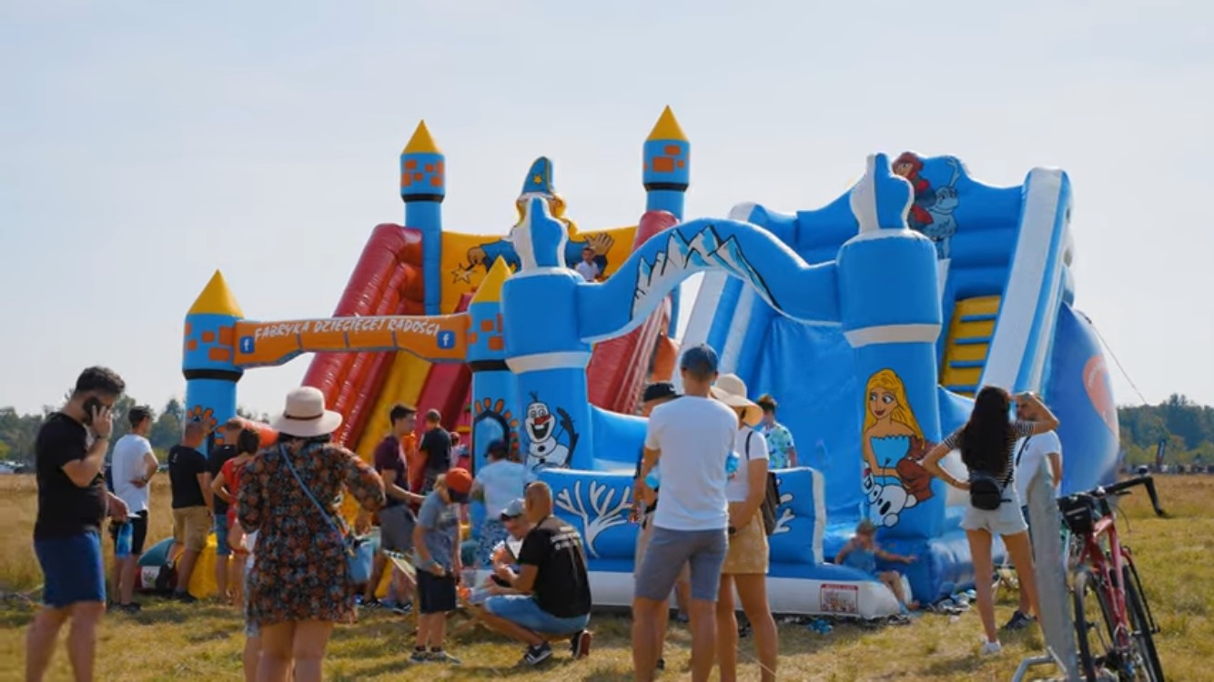 Kolorowy, dmuchany plac zabaw z motywami z bajki 'Kraina Lodu', otoczony grupą ludzi w słoneczny dzień, z rowerem zaparkowanym z boku.