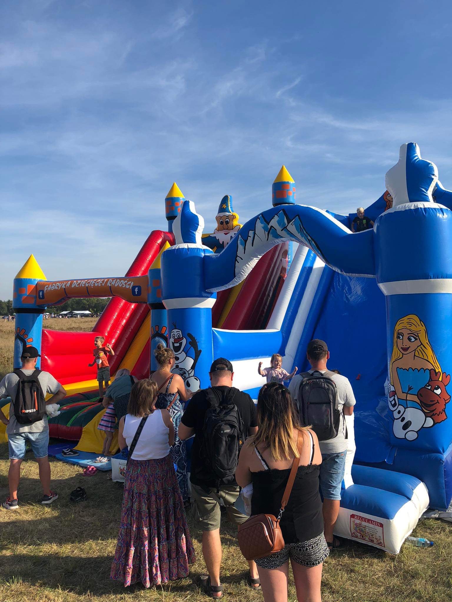 Duży, kolorowy dmuchany zamek z motywami bajkowymi, zjeżdżalnią i napisem 'Fabryka Dziecięcej Radości', otoczony grupą ludzi czekających w kolejce, na tle trawiastego terenu i błękitnego nieba.