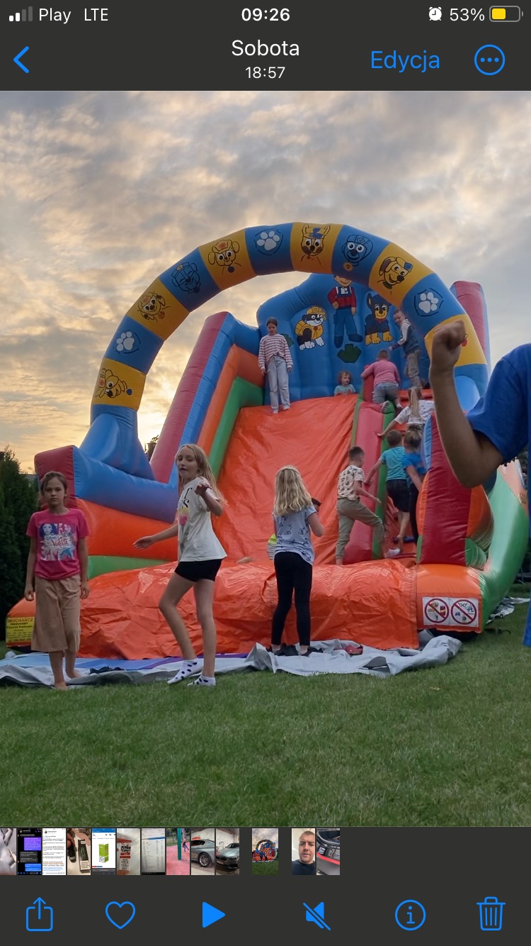 Dzieci bawią się na dmuchanej zjeżdżalni z motywem bajki 'Psi Patrol' w ogrodzie o zmierzchu.