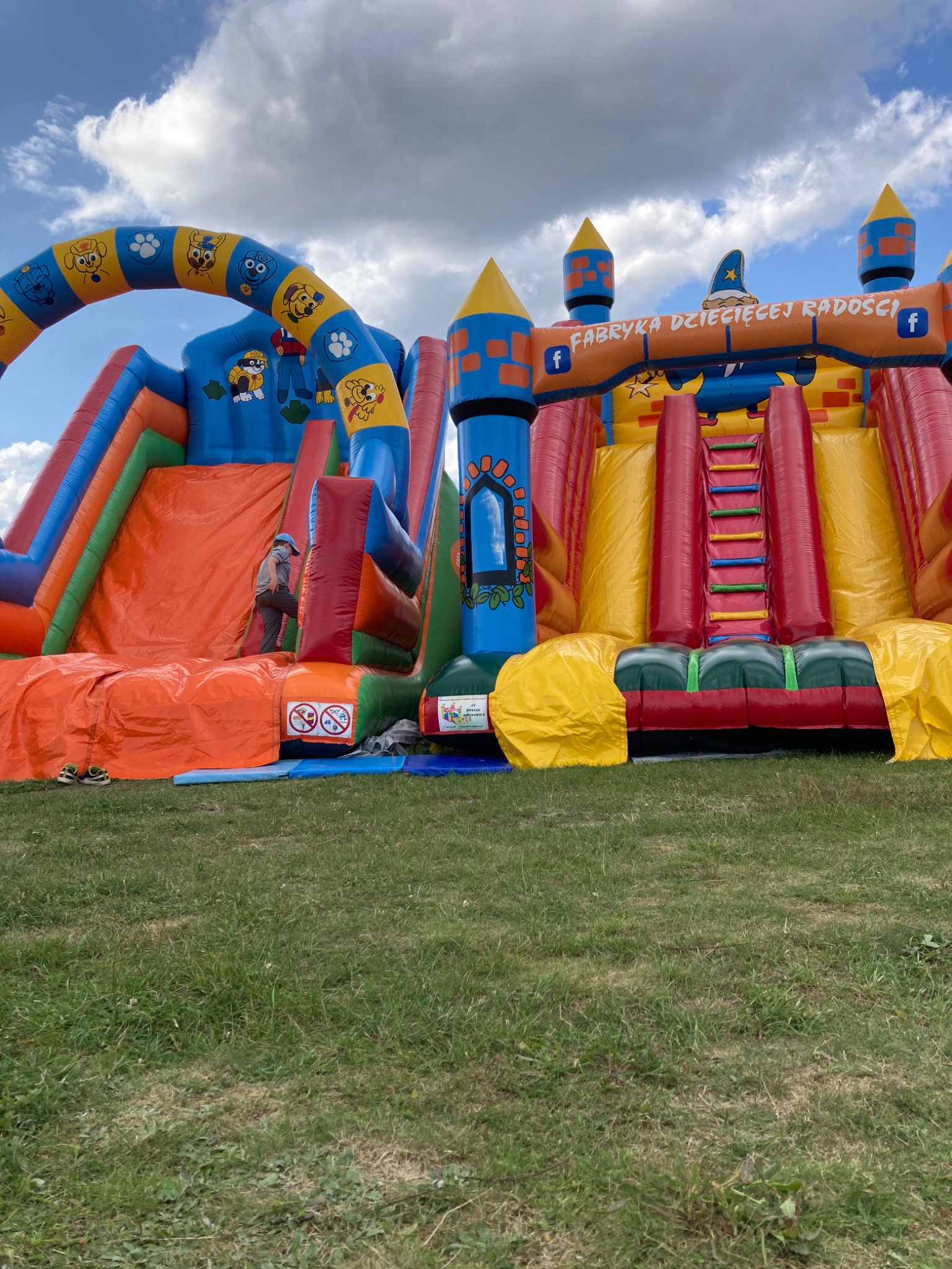 Kolorowy, dmuchany plac zabaw z motywami bajkowymi i napisem 'Fabryka Dziecięcej Radości', częściowo rozłożony na trawniku, z widoczną osobą wchodzącą na zjeżdżalnię i parą butów obok.