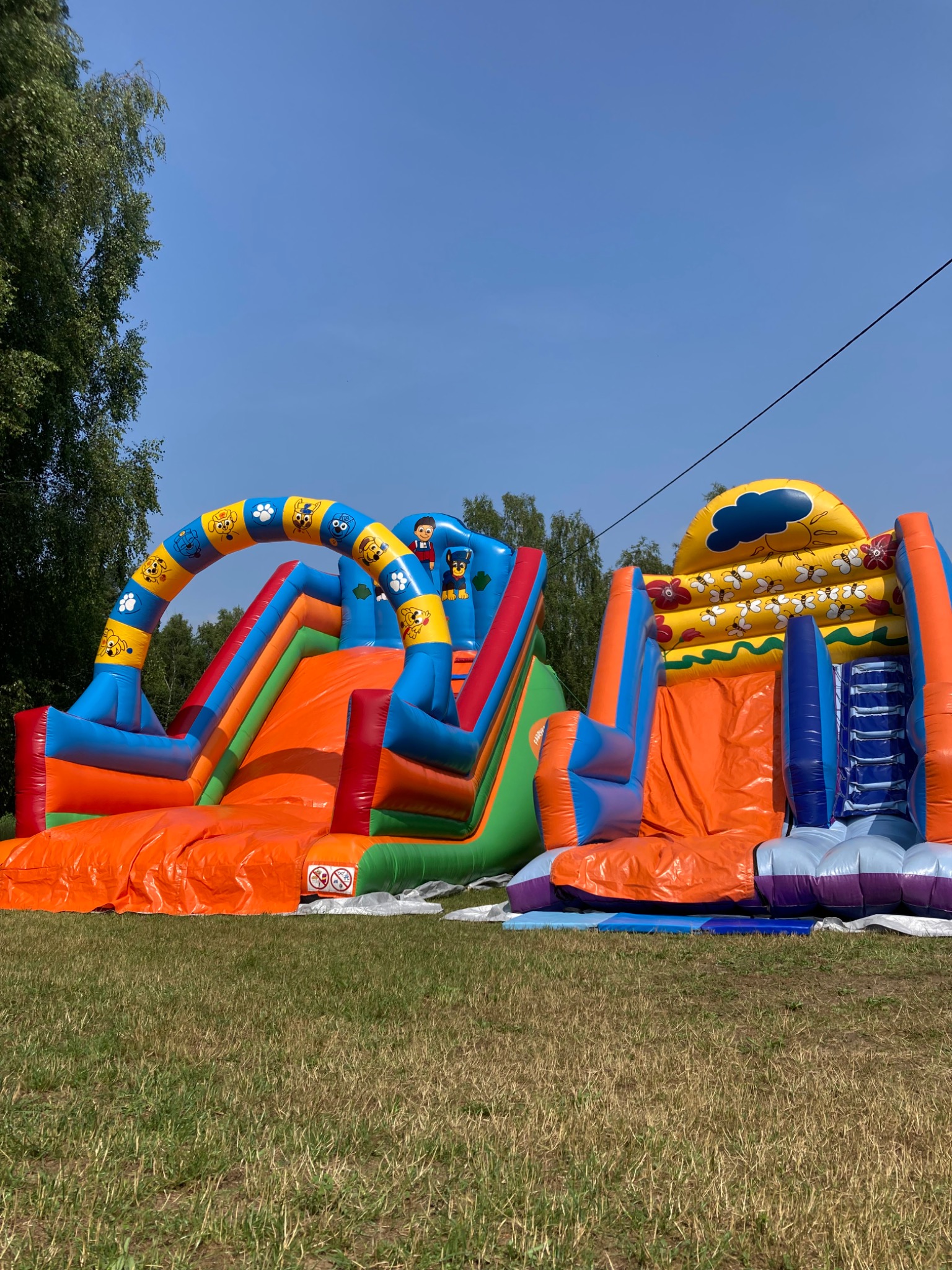 Dwa kolorowe, nadmuchiwane zamki ze zjeżdżalniami na trawniku w słoneczny dzień. Jeden z motywem psów, drugi z motywem słońca i chmur.