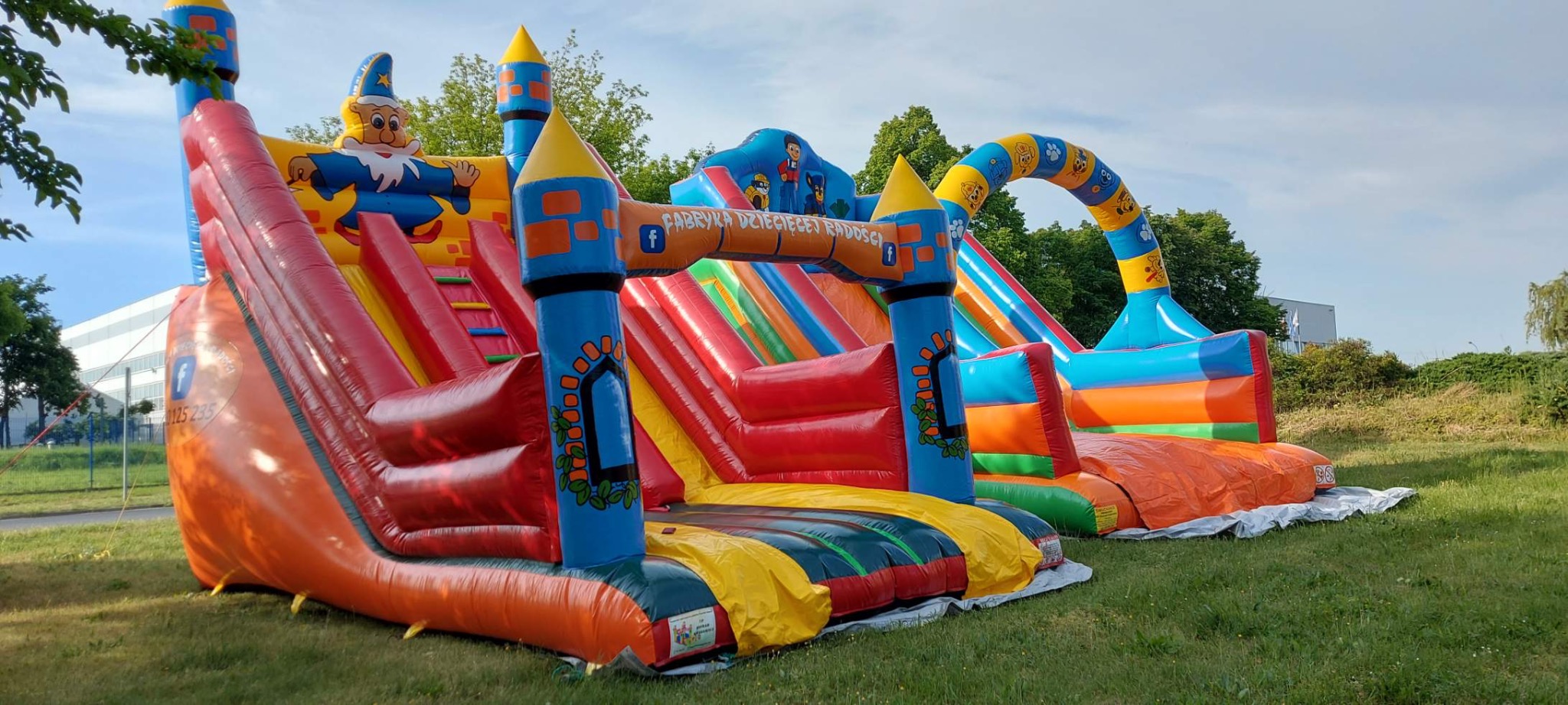 Kolorowy dmuchany zamek ze zjeżdżalniami, z motywami bajkowymi i logo firmy, ustawiony na trawie.