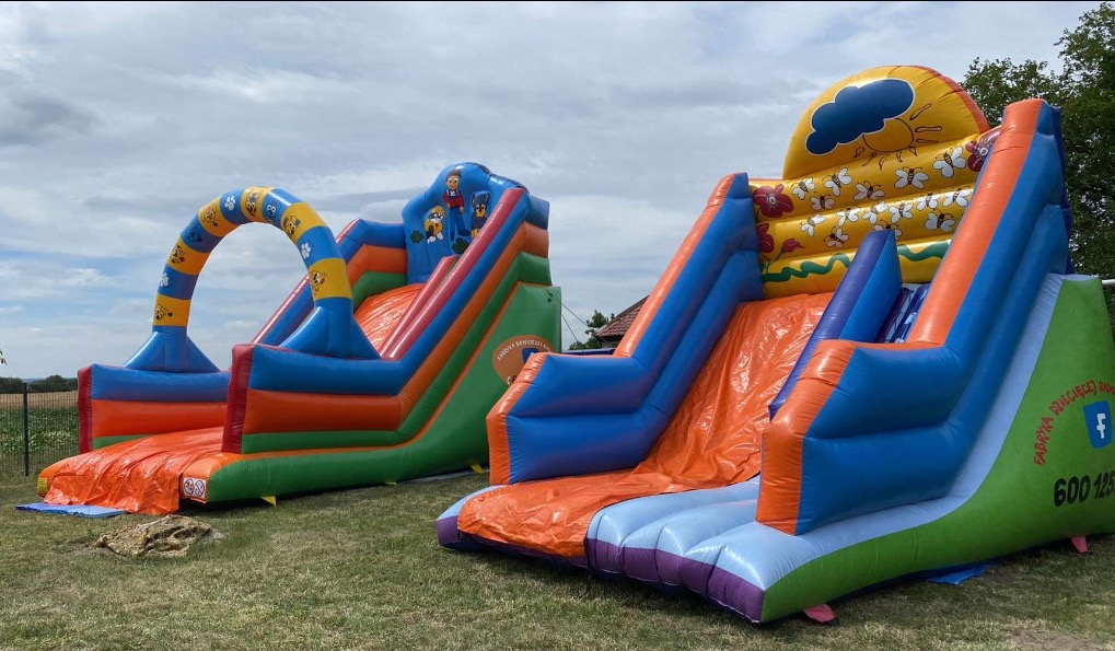 Dwa kolorowe dmuchane zamki ze zjeżdżalniami na trawie, jeden z motywem psiego patrolu, drugi z motywem słońca i pszczół, widoczny numer telefonu firmy na jednym z nich.