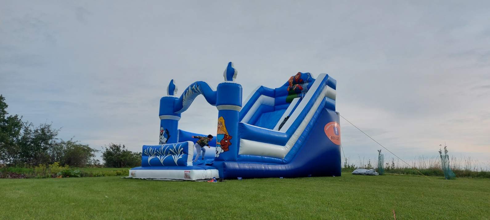 Niebiesko-biały dmuchany zamek ze zjeżdżalnią na trawie, z motywami postaci z bajek, dzieci bawiące się w środku.