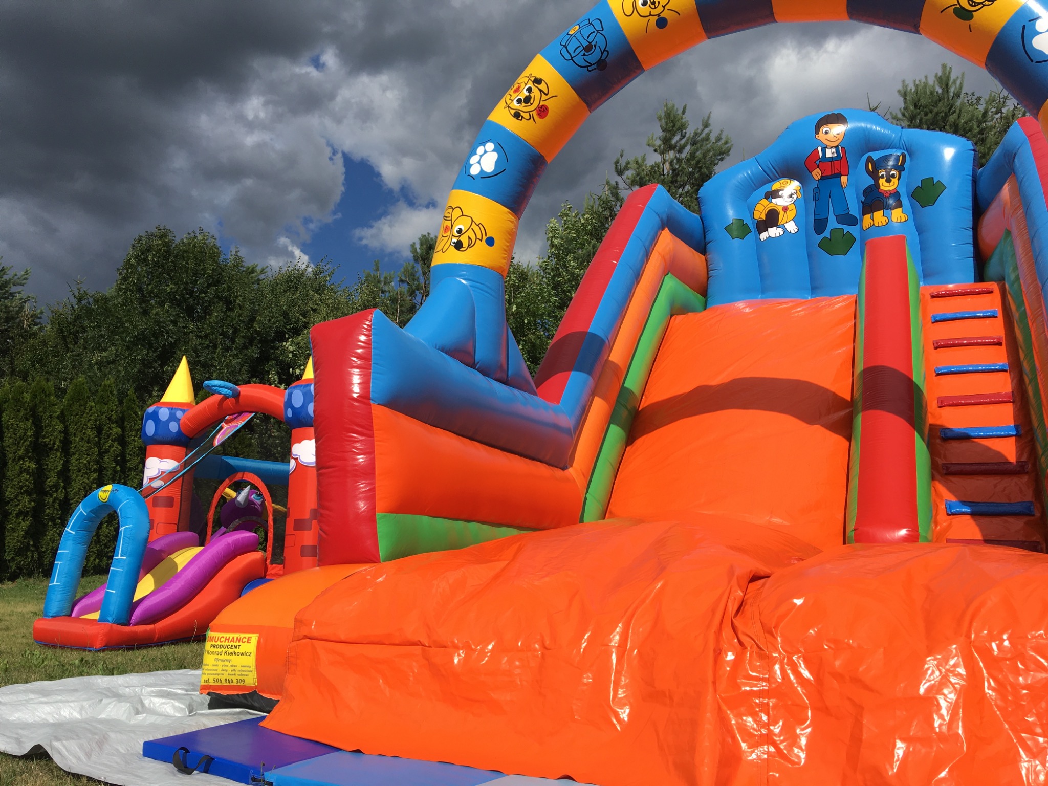 Kolorowy, dmuchany plac zabaw z motywami z bajki Psi Patrol na zielonej trawie, widoczny fragment tabliczki informacyjnej producenta.