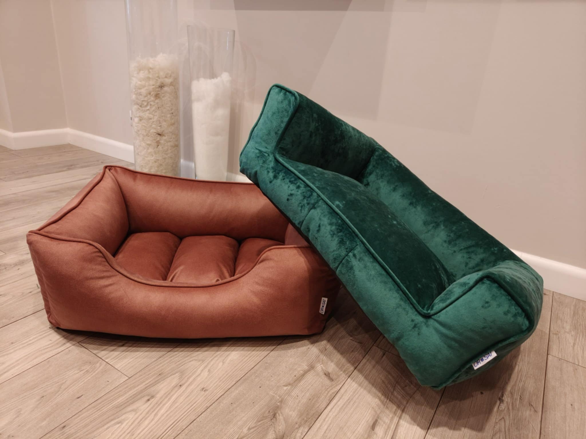 Dwa welurowe legowiska dla zwierząt: brązowe z poduszkami i zielone, ustawione na drewnianej podłodze, z pojemnikami z wypełnieniem w tle.