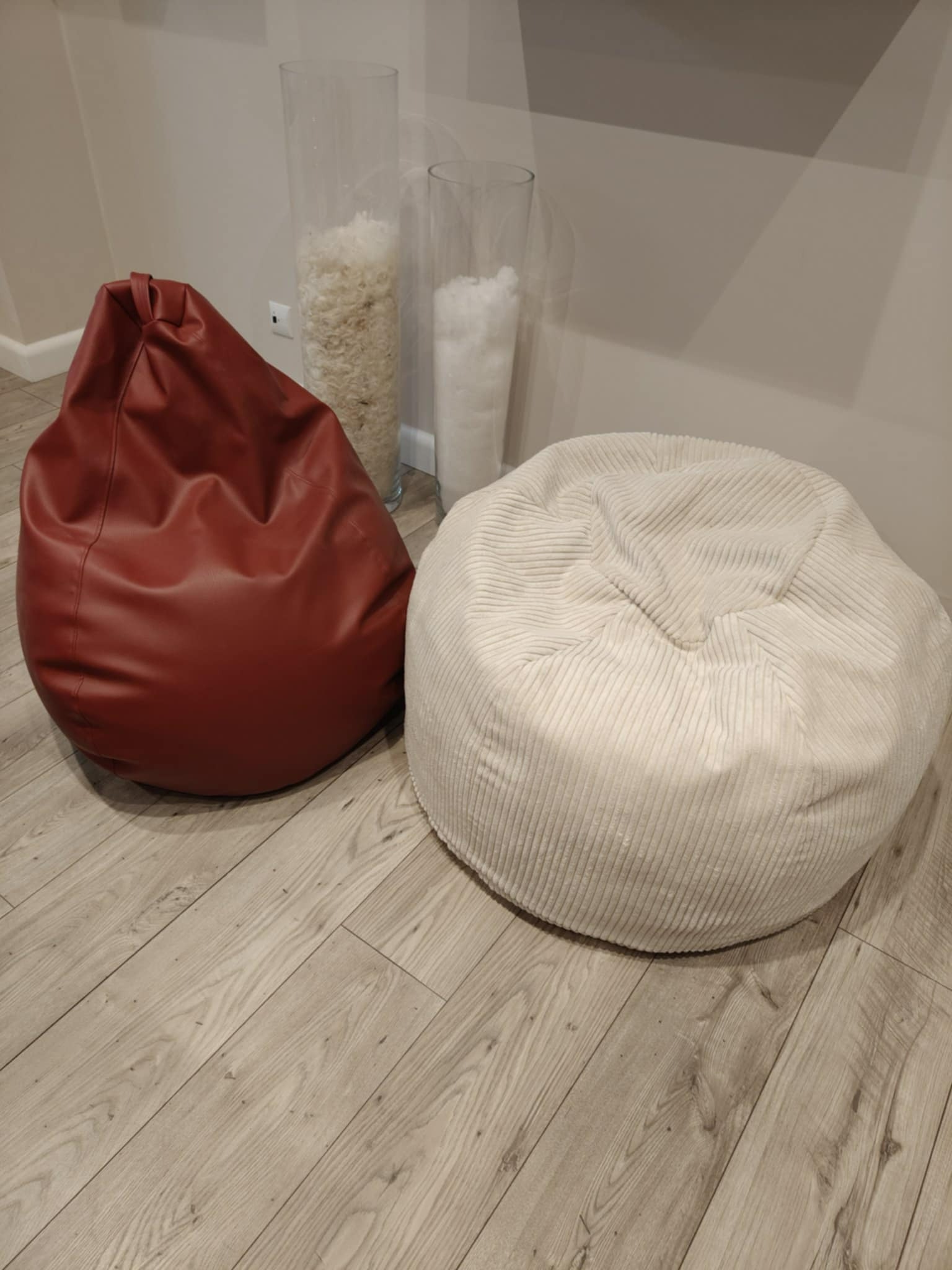 Dwa pufy: bordowy skórzany i biały z prążkowanej tkaniny, stoją na drewnianej podłodze obok szklanych wazonów z białym wypełnieniem.