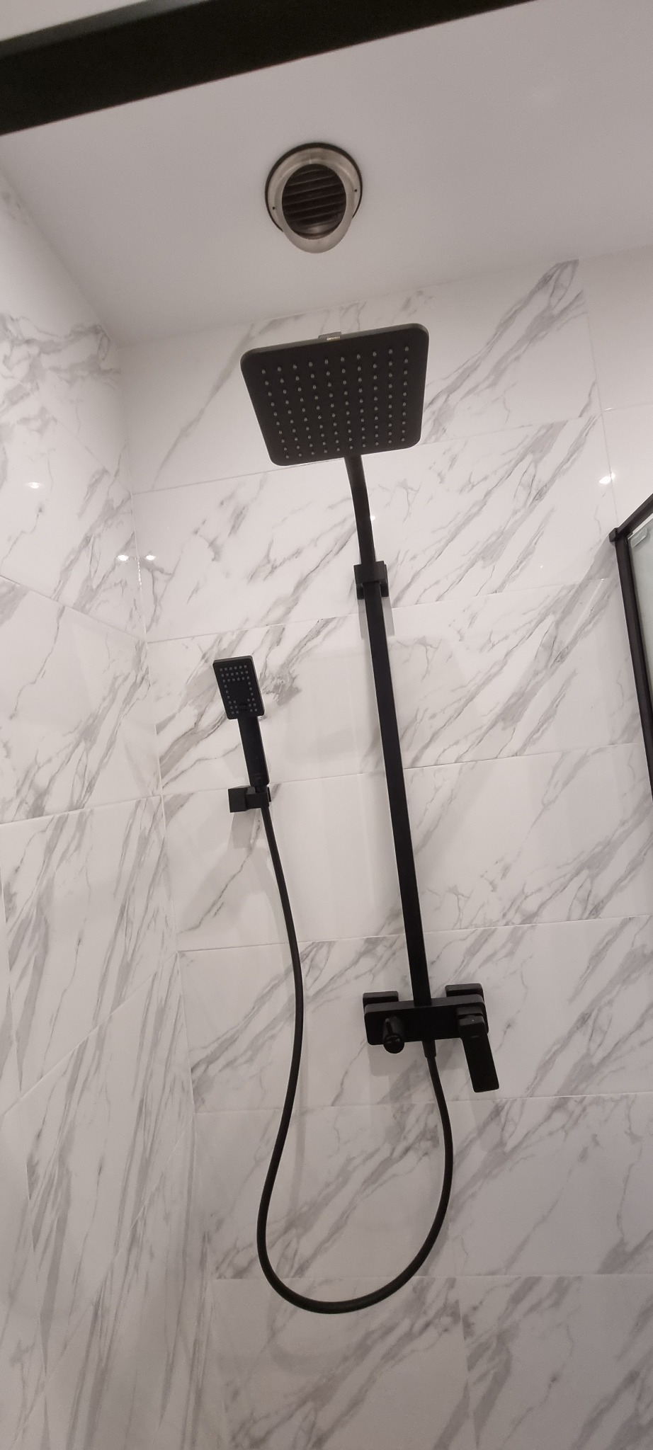 Czarna, kwadratowa deszczownica i słuchawka prysznicowa zamontowane na ścianie z białych płytek imitujących marmur w wykończonej łazience.
