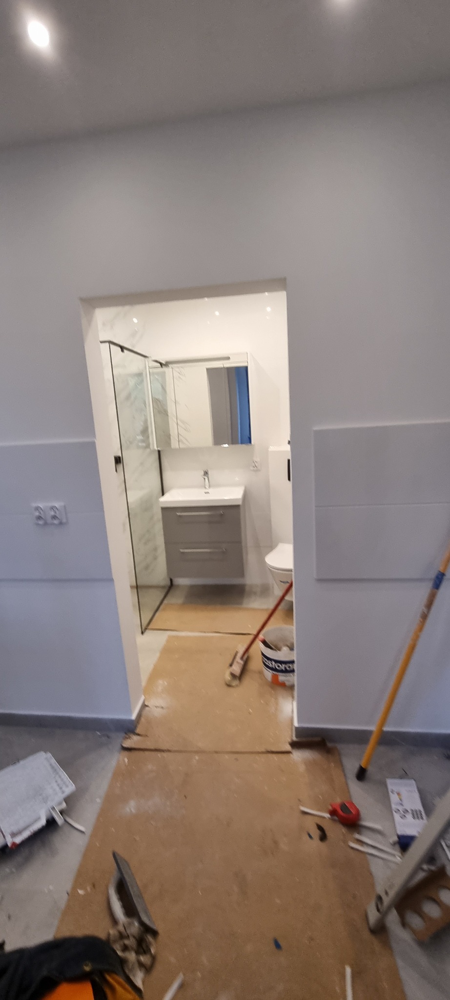 Remont łazienki w Szczecinie: Widok na częściowo wykończoną łazienkę z kabiną prysznicową, umywalką z szafką i toaletą. Podłoga zabezpieczona kartonem, widoczne narzędzia i wiadro z farbą.