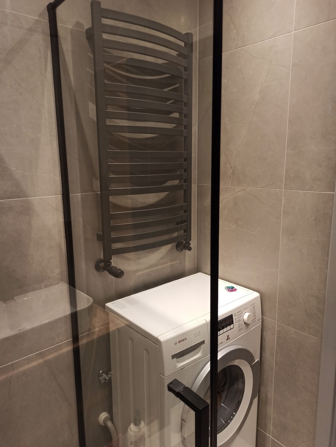 Szara łazienka z płytkami imitującymi marmur, grzejnik drabinkowy w kolorze grafitowym i biała pralka Bosch Serie 4, widok zza szklanej kabiny prysznicowej.