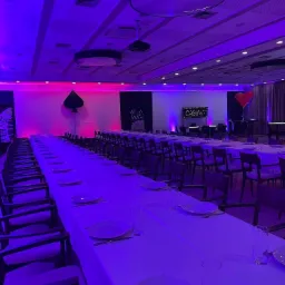 Długi stół bankietowy nakryty białym obrusem, z zastawą i szklankami, oświetlony fioletowym światłem, w tle dekoracje tematyczne kasyna: pik i serce oraz podświetlany napis CASINO.