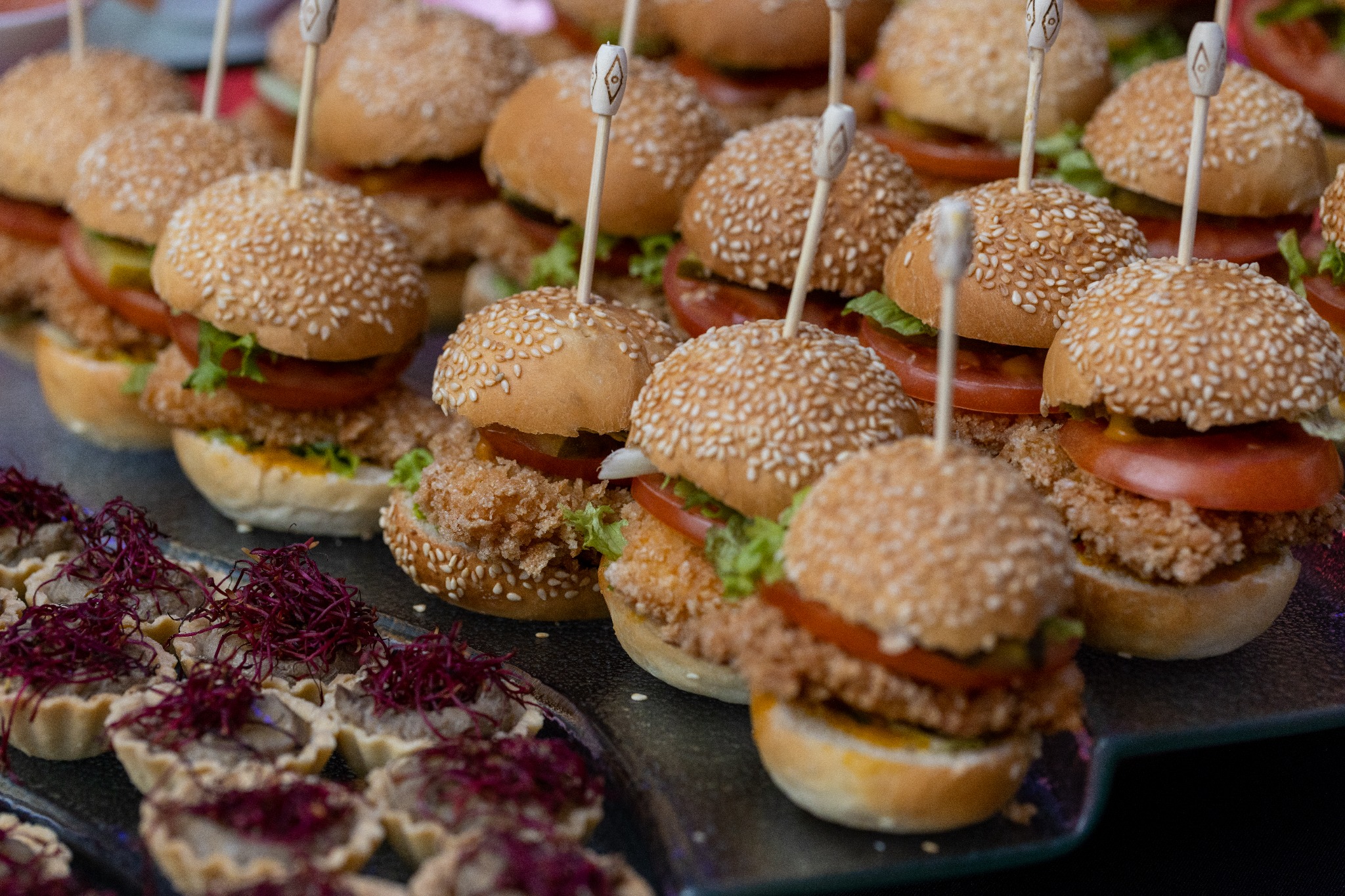 Miniaturowe burgery z panierowanym mięsem, pomidorem i sałatą, spięte dekoracyjnymi wykałaczkami, oraz tartaletki z ciemnym farszem i dekoracją z czerwonych pędów na ciemnym szklanym półmisku.