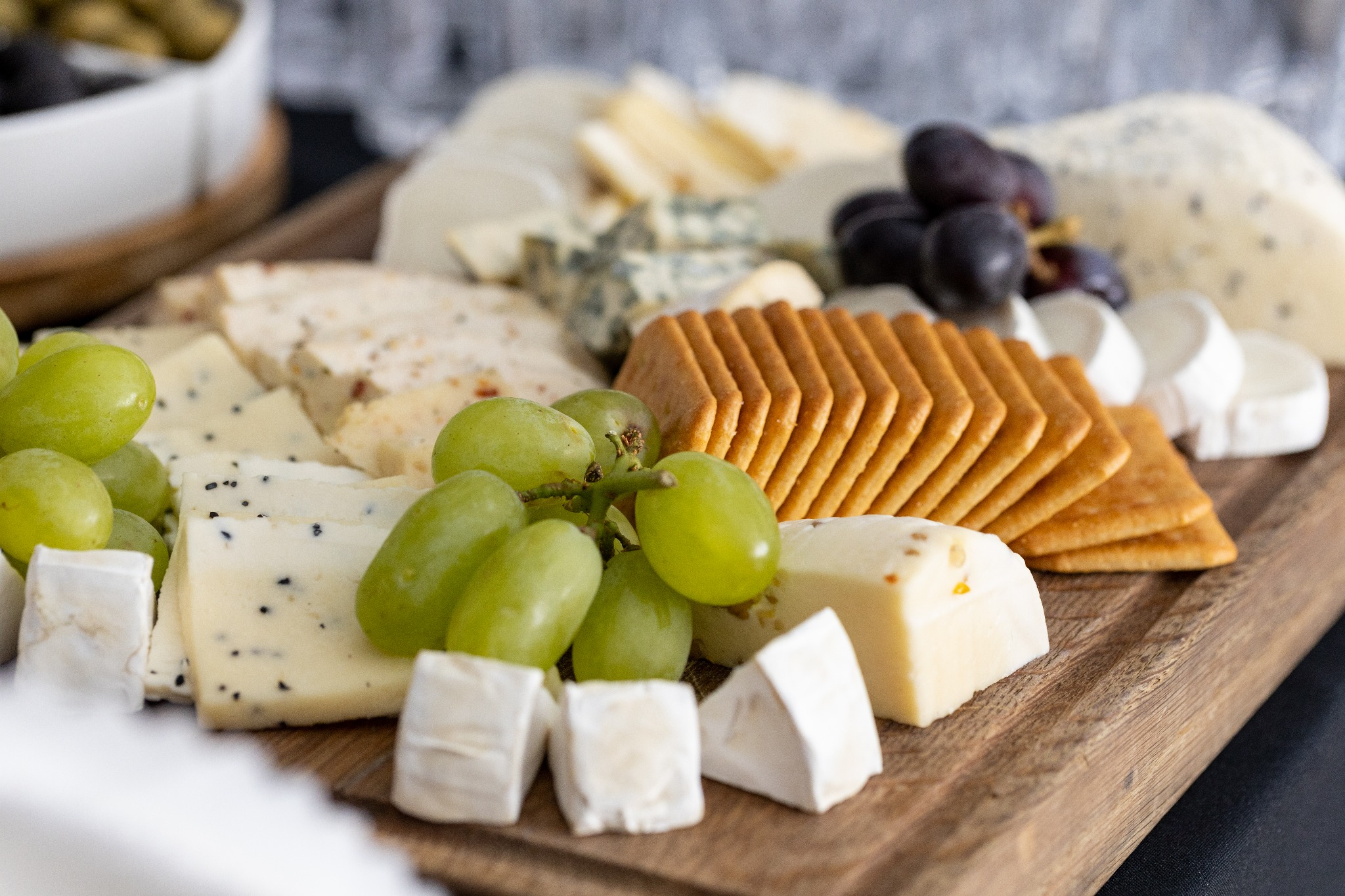 Deska serów z różnymi gatunkami, krakersami i owocami (winogrona zielone i ciemne), ułożona na drewnianej tacy, z białym naczyniem z oliwkami w tle.
