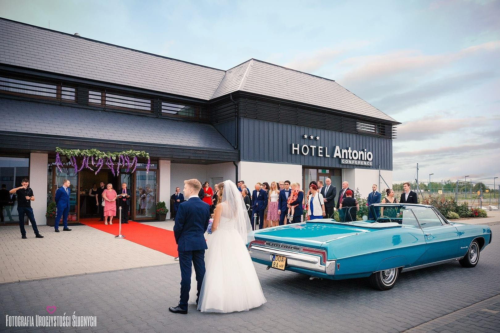 Para młoda przed hotelem Antonio Conference z turkusowym kabrioletem w tle, goście weselni na czerwonym dywanie przed wejściem.