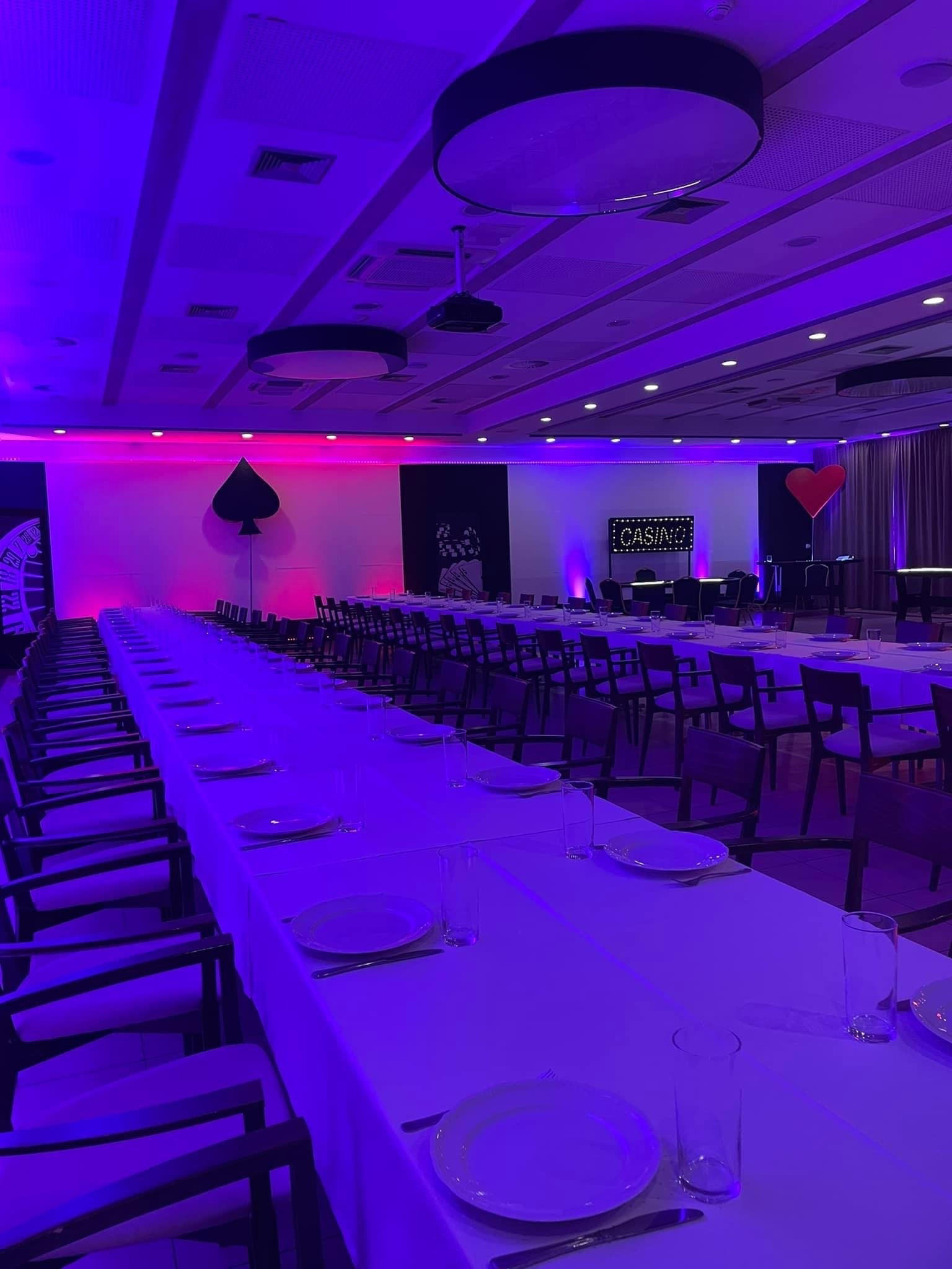 Długi stół bankietowy nakryty białym obrusem, z zastawą i szklankami, oświetlony fioletowym światłem, w tle dekoracje tematyczne kasyna: pik i serce oraz podświetlany napis CASINO.