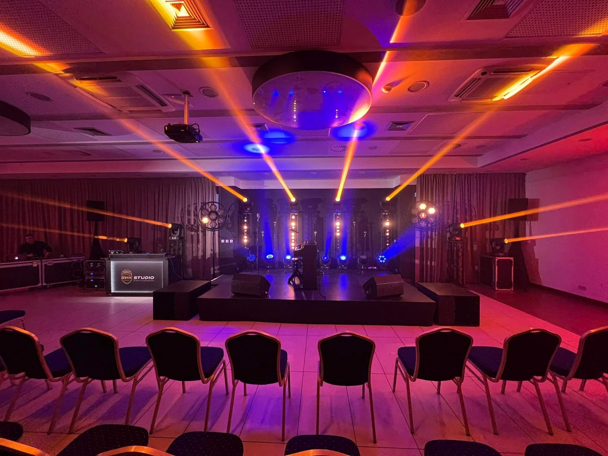 Sala konferencyjna przygotowana na wydarzenie z oświetleniem scenicznym, podium i rzędami krzeseł, widoczny sprzęt DJ-ski z logo SWK Studio
