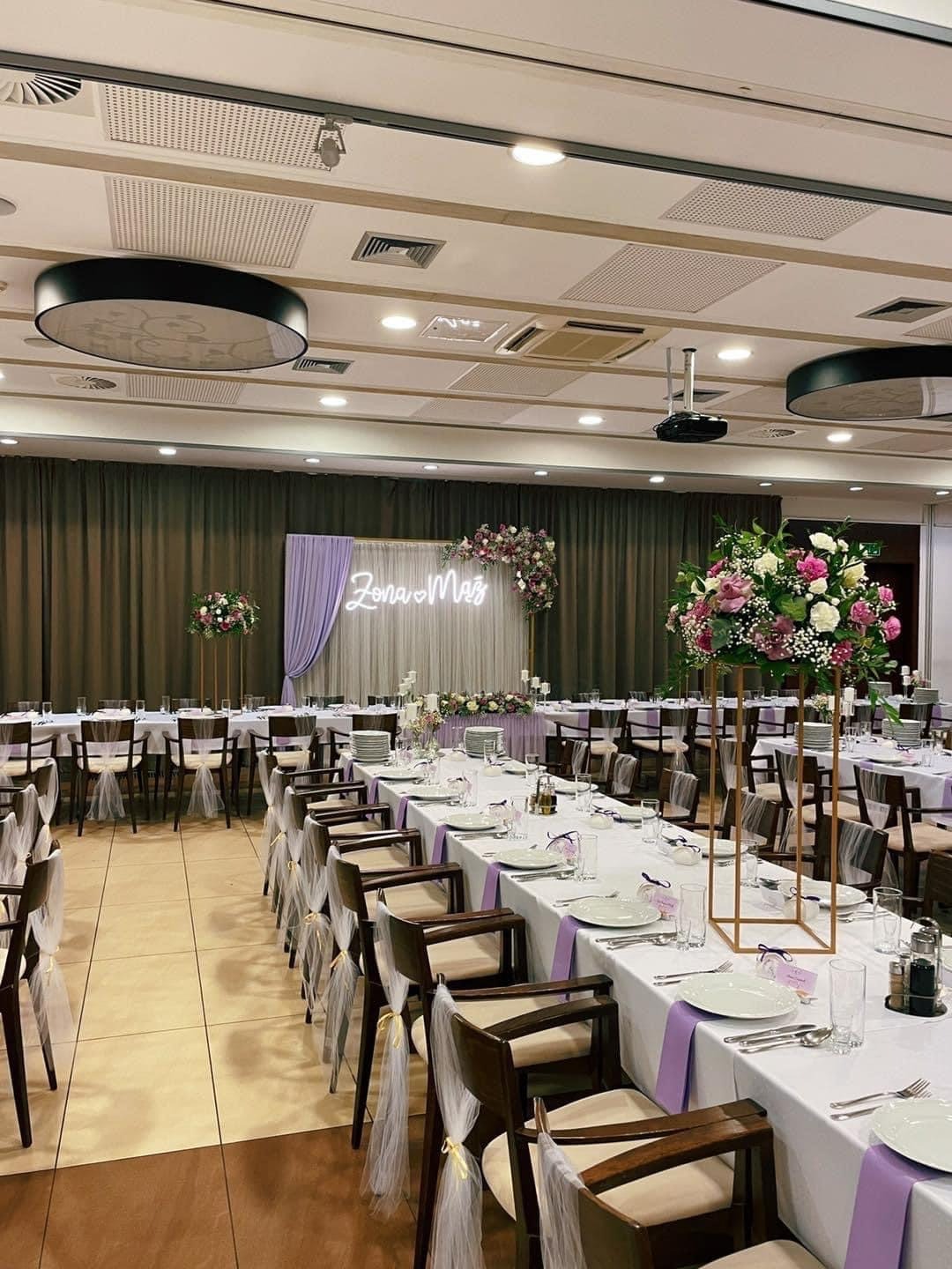 Uroczysta sala bankietowa z długimi stołami nakrytymi białymi obrusami, fioletowe akcenty, krzesła z tiulowymi ozdobami, dekoracje kwiatowe i neonowy napis 'Żona&Mąż' na tle zasłon.
