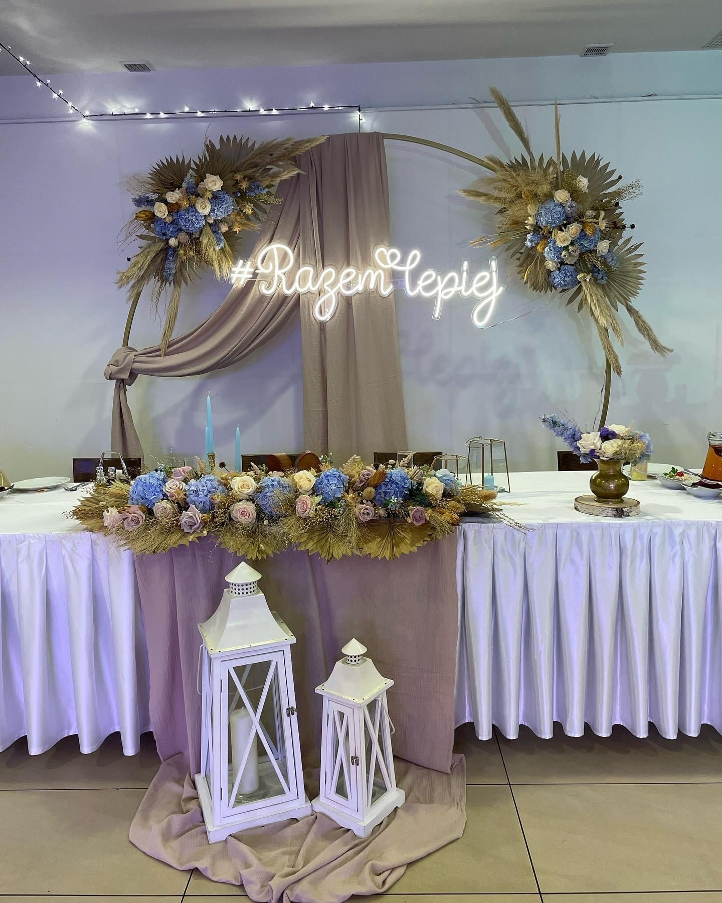 Udekorowany stół weselny z kwiatami, suszem, lampionami i neonowym napisem '#RazemLpiej' na tle jasnej ściany.