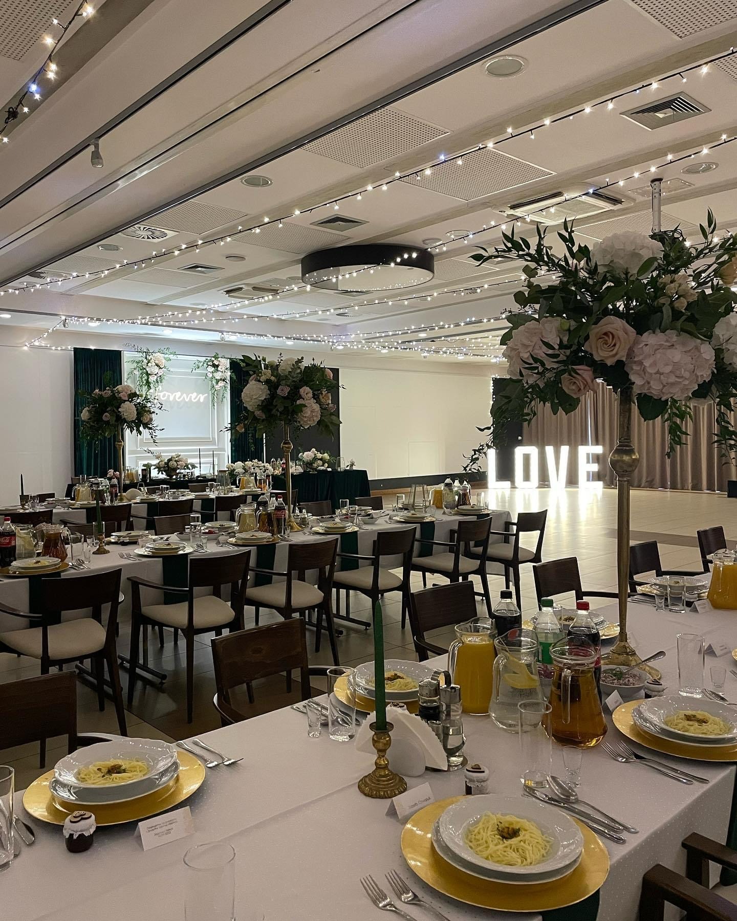 Uroczysta sala bankietowa z nakrytymi stołami, dekoracjami kwiatowymi, świecami w złotych świecznikach i podświetlanym napisem LOVE w tle.