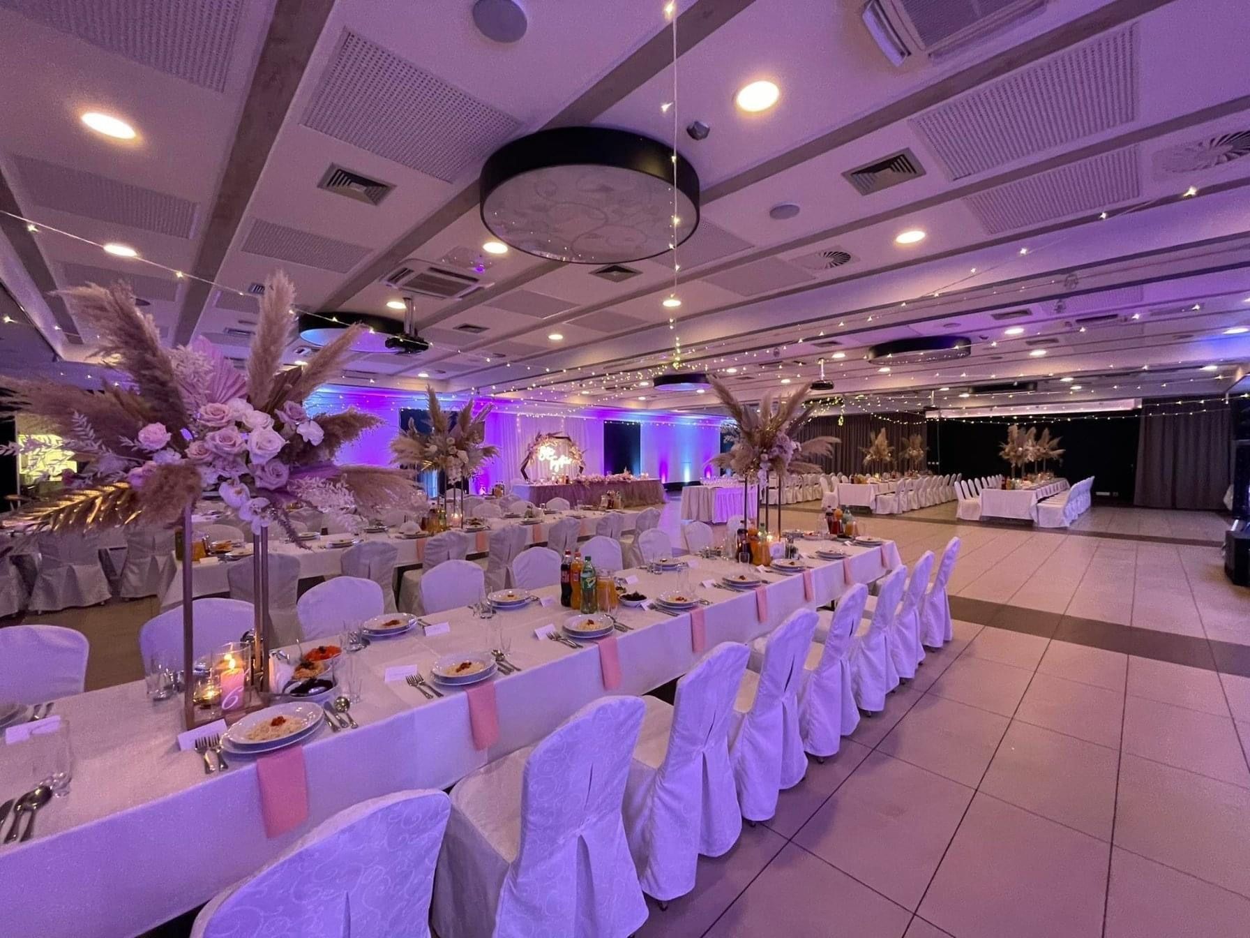 Uroczysta sala bankietowa przygotowana na przyjęcie, z długimi stołami nakrytymi białymi obrusami i krzesłami z białymi pokrowcami, ozdobiona kompozycjami kwiatowymi z róż i traw pampasowych...