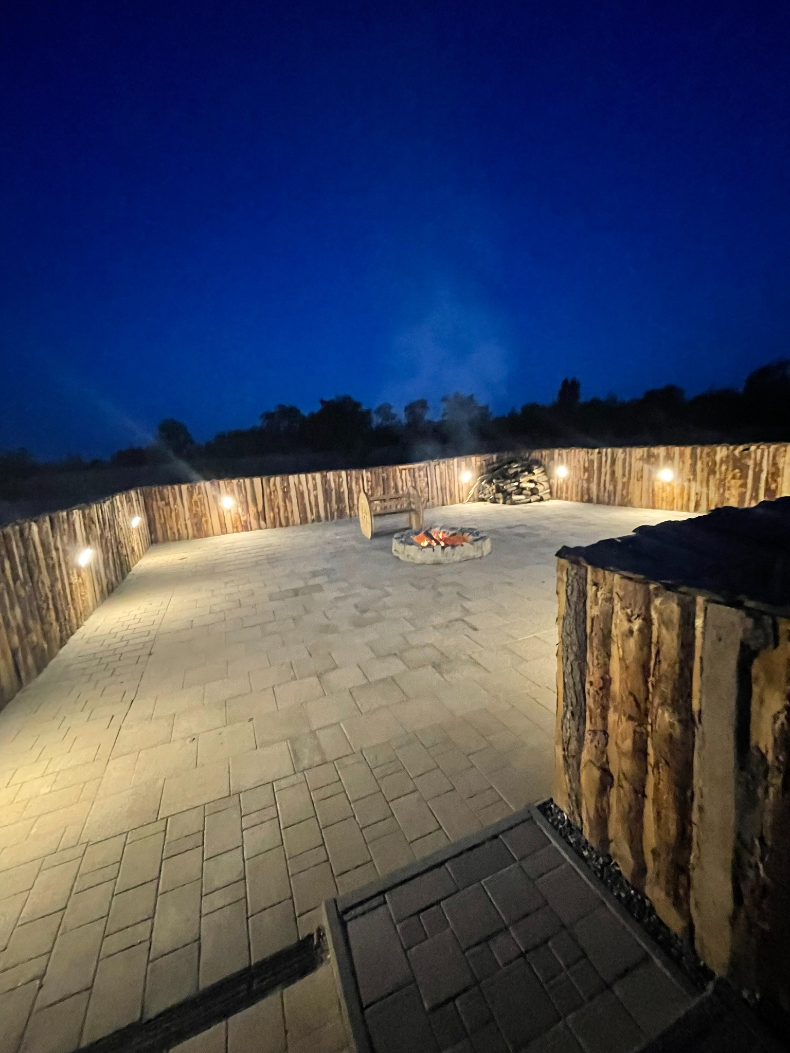 Oświetlona przestrzeń z paleniskiem otoczona drewnianym ogrodzeniem i brukowaną nawierzchnią, widok nocą.