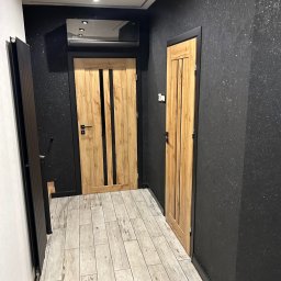 Multitechnika Piotr Koch Sp. z o.o. - Korytarz po remoncie: drewniane drzwi z czarnymi akcentami, czarne ściany z delikatnym brokatem, podłoga wyłożona płytkami imitującymi drewno, nowoczesne oświetlenie sufitowe.