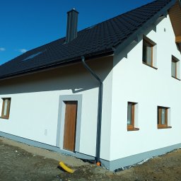 Multitechnika Piotr Koch Sp. z o.o. - Nowoczesny dom jednorodzinny z białą elewacją i ciemną dachówką, widok z boku na świeżo wykończoną ścianę z brązowymi oknami i drzwiami, żółta rura drenażowa leżąca obok fundamentu.