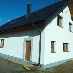 Nowoczesny dom jednorodzinny z białą elewacją i ciemną dachówką, widok z boku na świeżo wykończoną ścianę z brązowymi oknami i drzwiami, żółta rura drenażowa leżąca obok fundamentu.