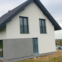 Multitechnika Piotr Koch Sp. z o.o. - Nowoczesny dom jednorodzinny z białą elewacją i szarym cokołem, okna zabezpieczone folią, widoczny fragment ogrodu i krajobraz w tle.