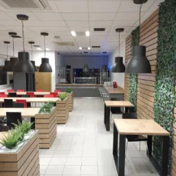 Nowoczesna aranżacja wnętrza biurowej stołówki z drewnianymi panelami ściennymi, roślinami w donicach i wiszącymi lampami w stylu industrialnym.