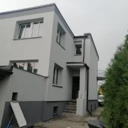 Dwukondygnacyjny dom po odnowieniu elewacji, pomalowany w odcieniach szarości i bieli, z widocznymi śladami prac remontowych na podwórku.