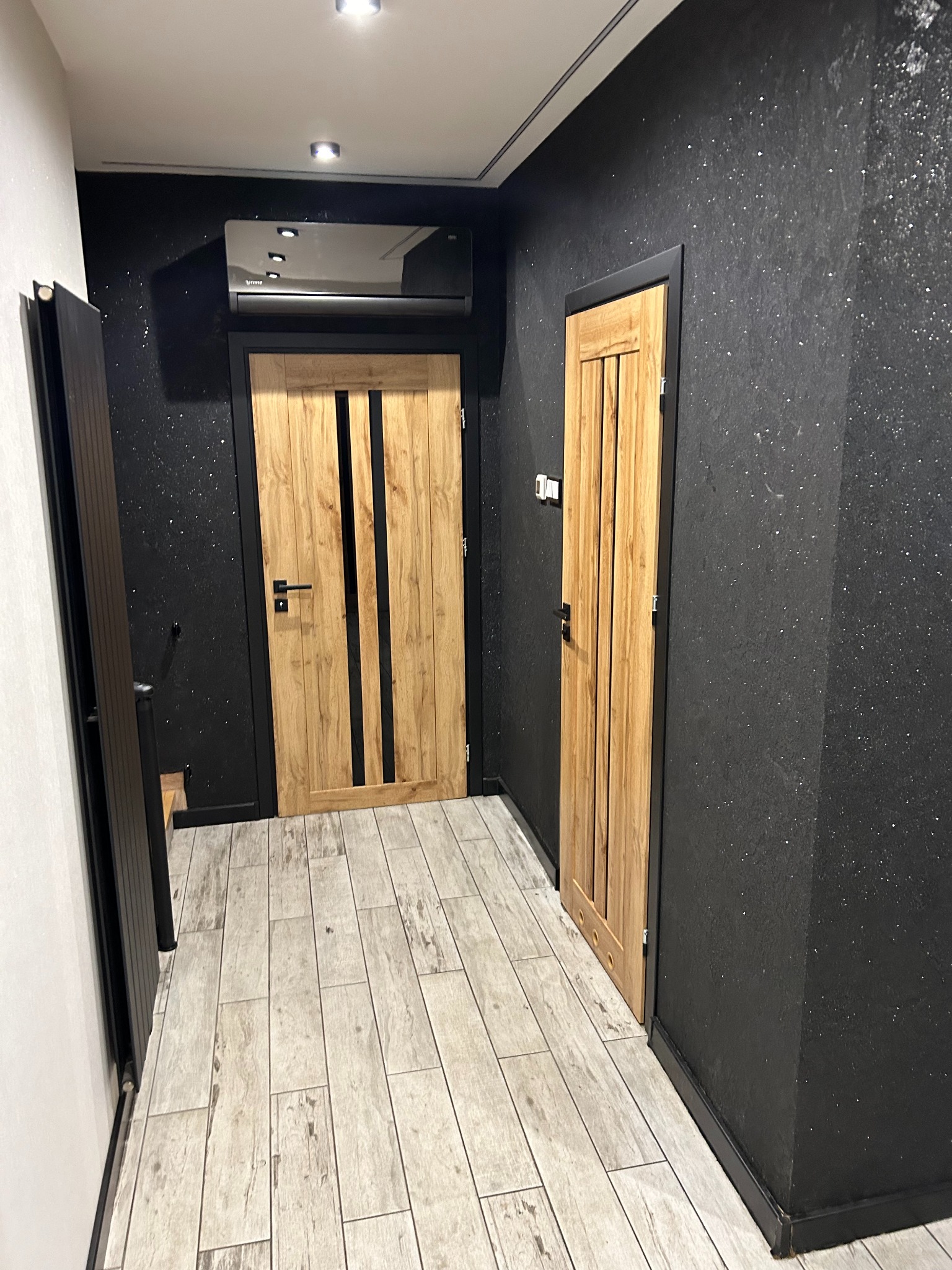 Korytarz po remoncie: drewniane drzwi z czarnymi akcentami, czarne ściany z delikatnym brokatem, podłoga wyłożona płytkami imitującymi drewno, nowoczesne oświetlenie sufitowe.